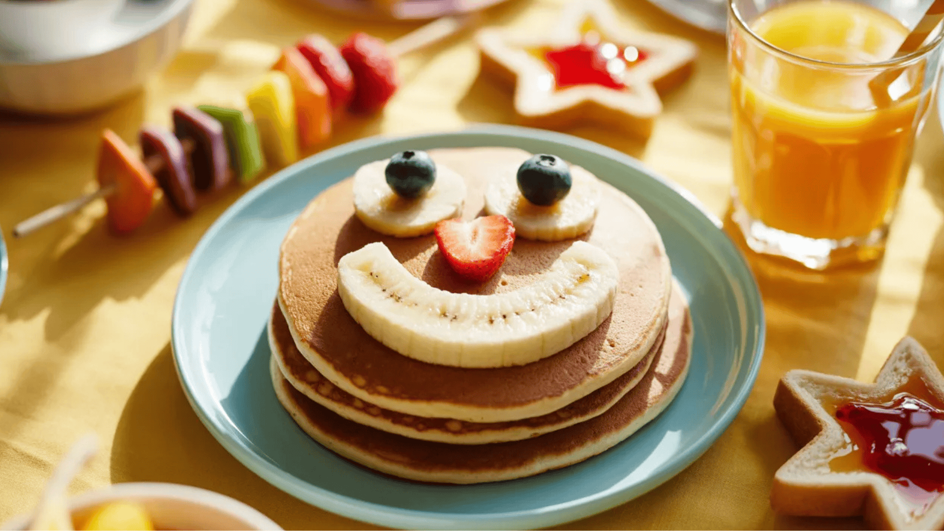 stack of pancakes topped with banana slices, strawberries, and blueberries arranged as a smiling face, served with juice and fruit sides