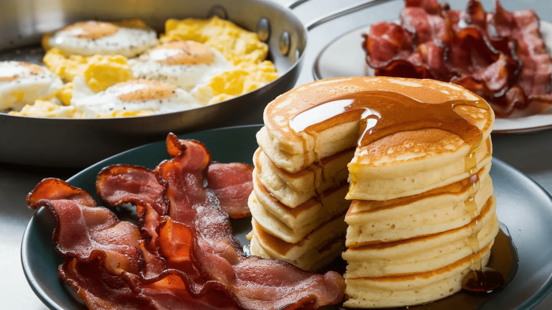 stack of pancakes topped with syrup served with crispy bacon and fried eggs in a skillet on a breakfast table