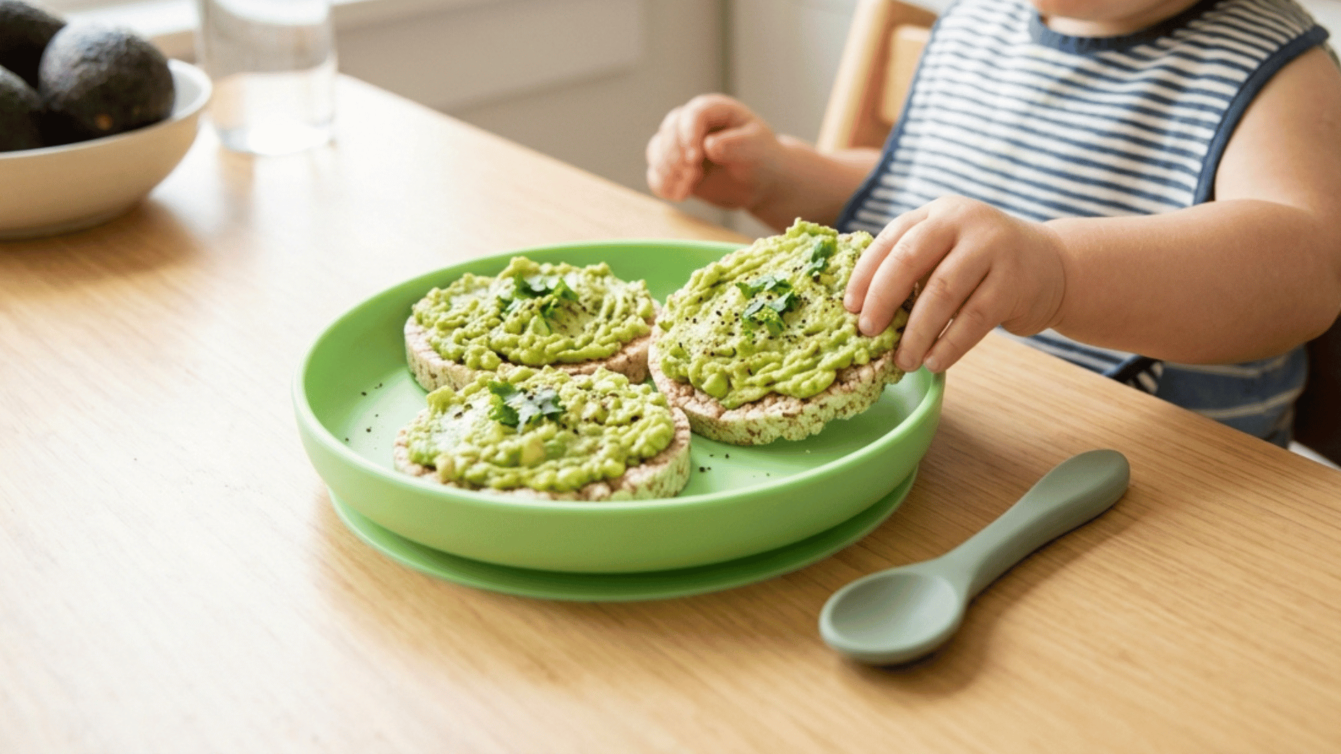 toddler eating rice cakes topped with mashed avocado on a green plate.