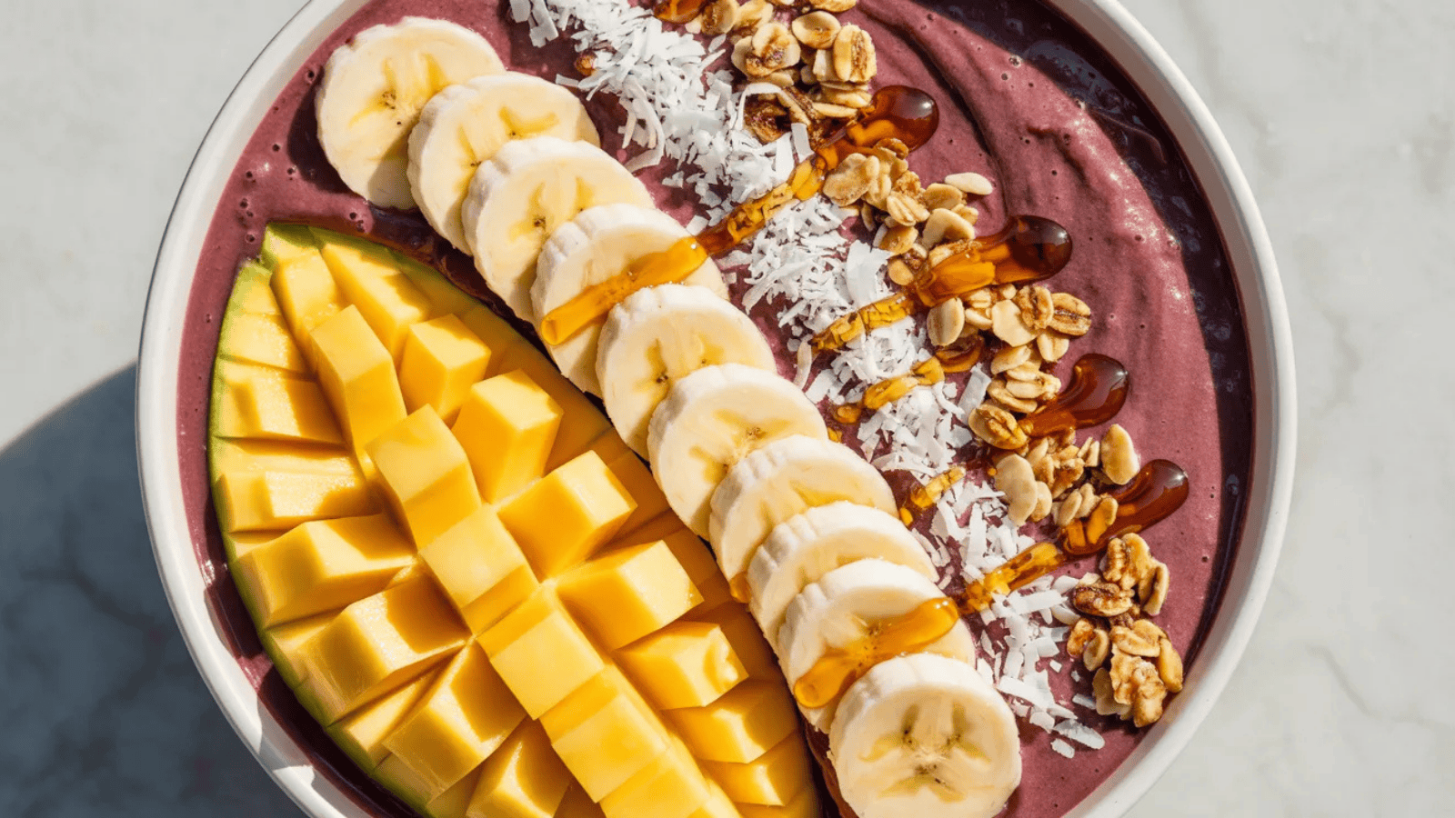 wide bowl filled with a deep purple acai smoothie base, topped with neat slices of fresh mango, banana rounds, shredded coconut
