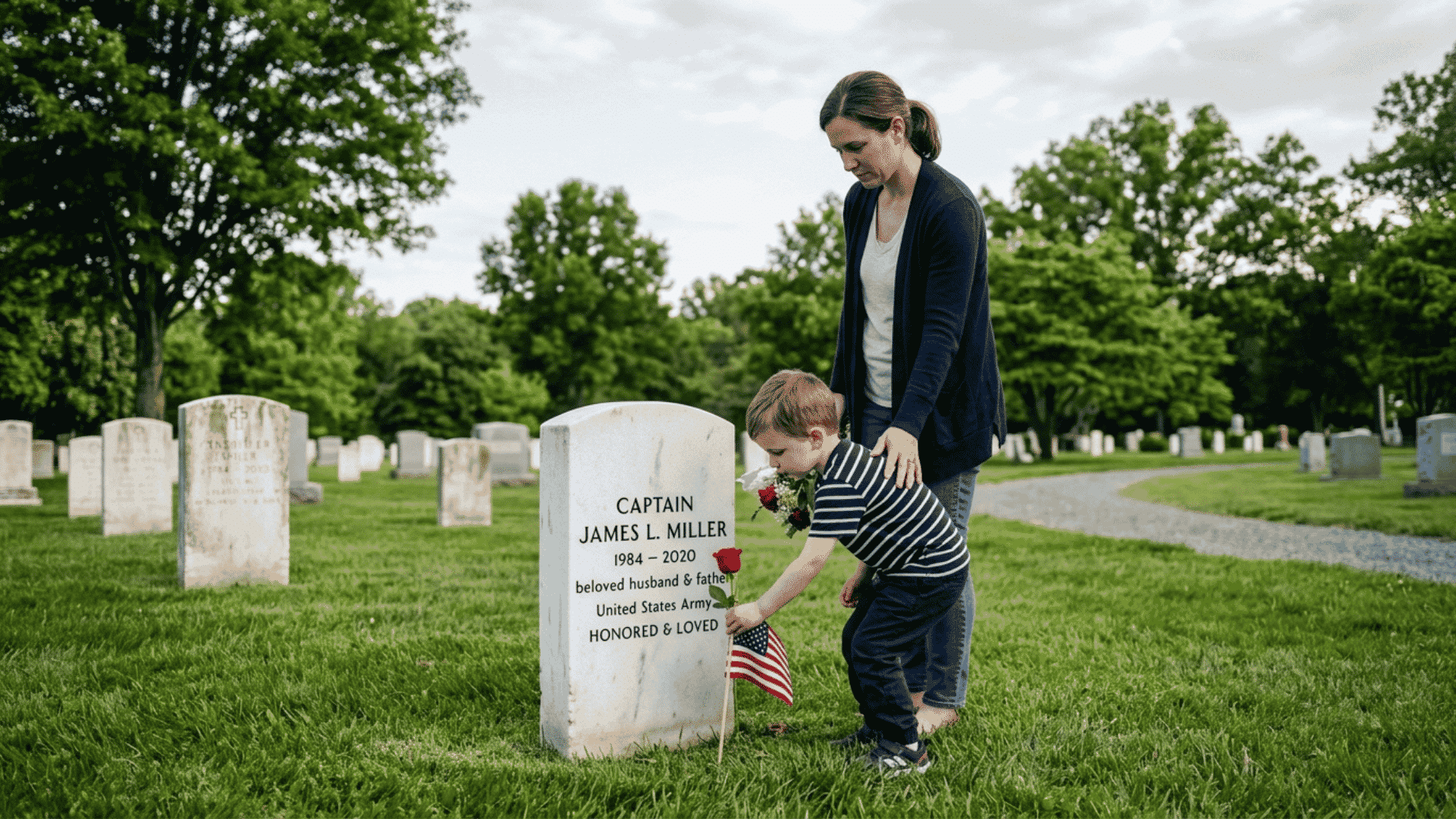 a parent and young child placing a red rose and small american flag at a white stone grave marker at a military cemetery on memorial day