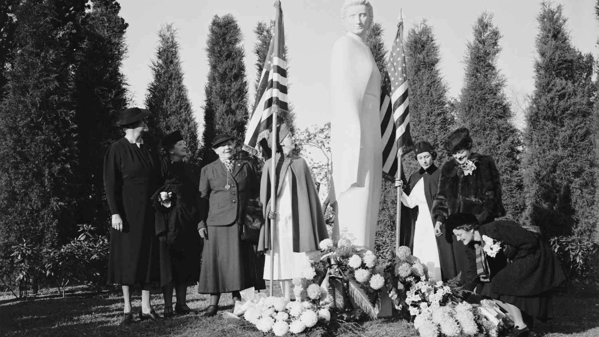 Black-and-white photo of women placing flowers at a memorial statue with American flags during a remembrance ceremony