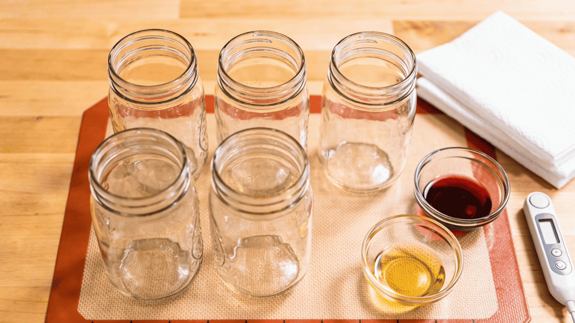 candle making workspace with mason jars, silicone mat, fragrance oils and thermometer arranged on a wooden counter.