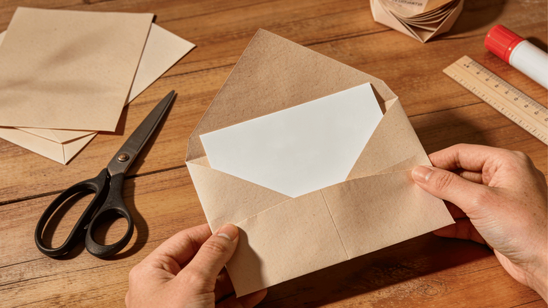 hands holding handmade kraft paper envelope with blank card on wooden table beside scissors and craft supplies