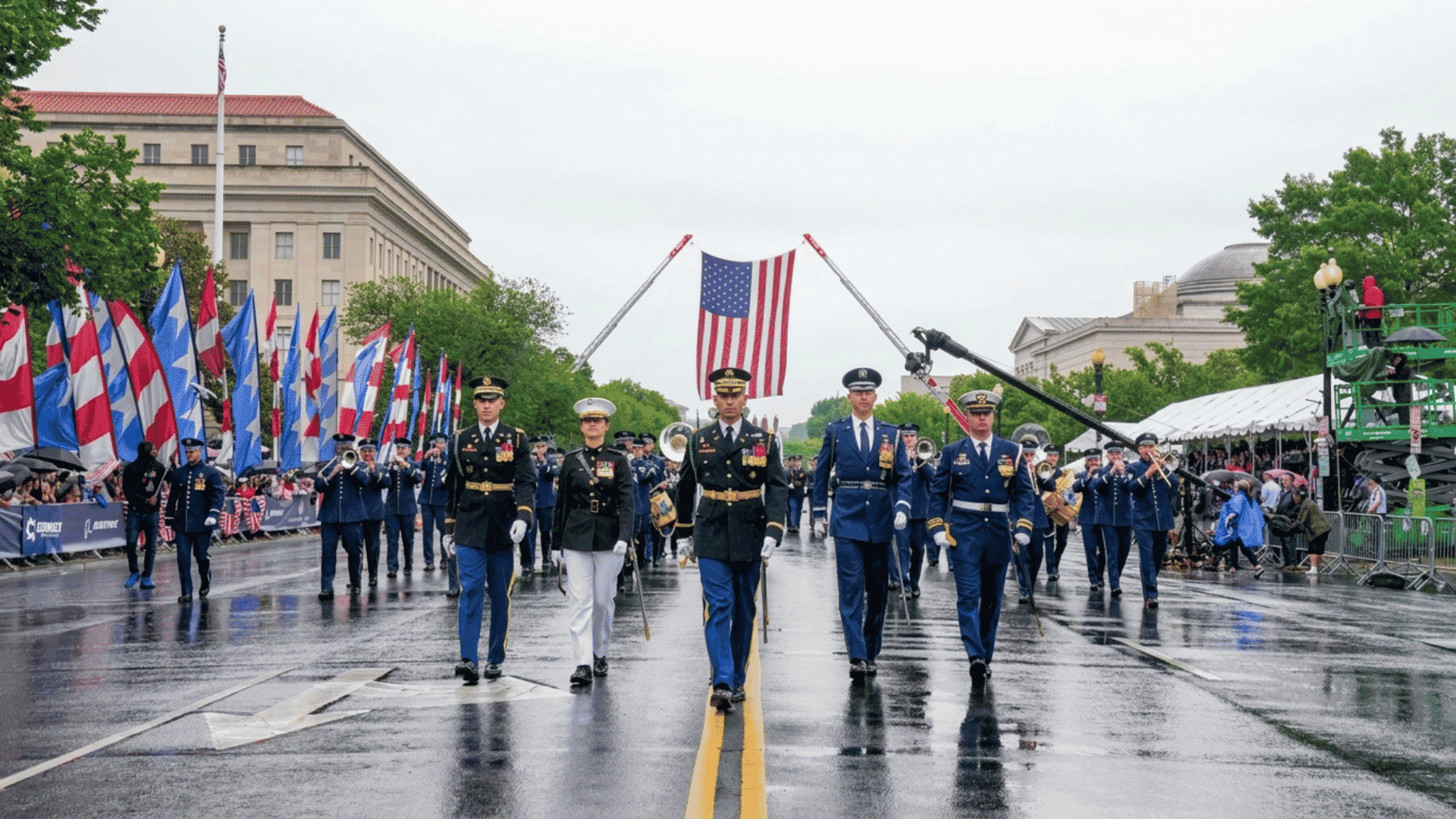 military bands and service members march down a rain-soaked street beneath a large american flag