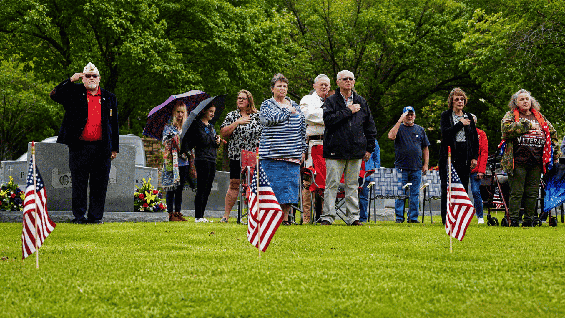 people stand in tribute during a memorial day ceremony at a cemetery with american flags placed across the lawn