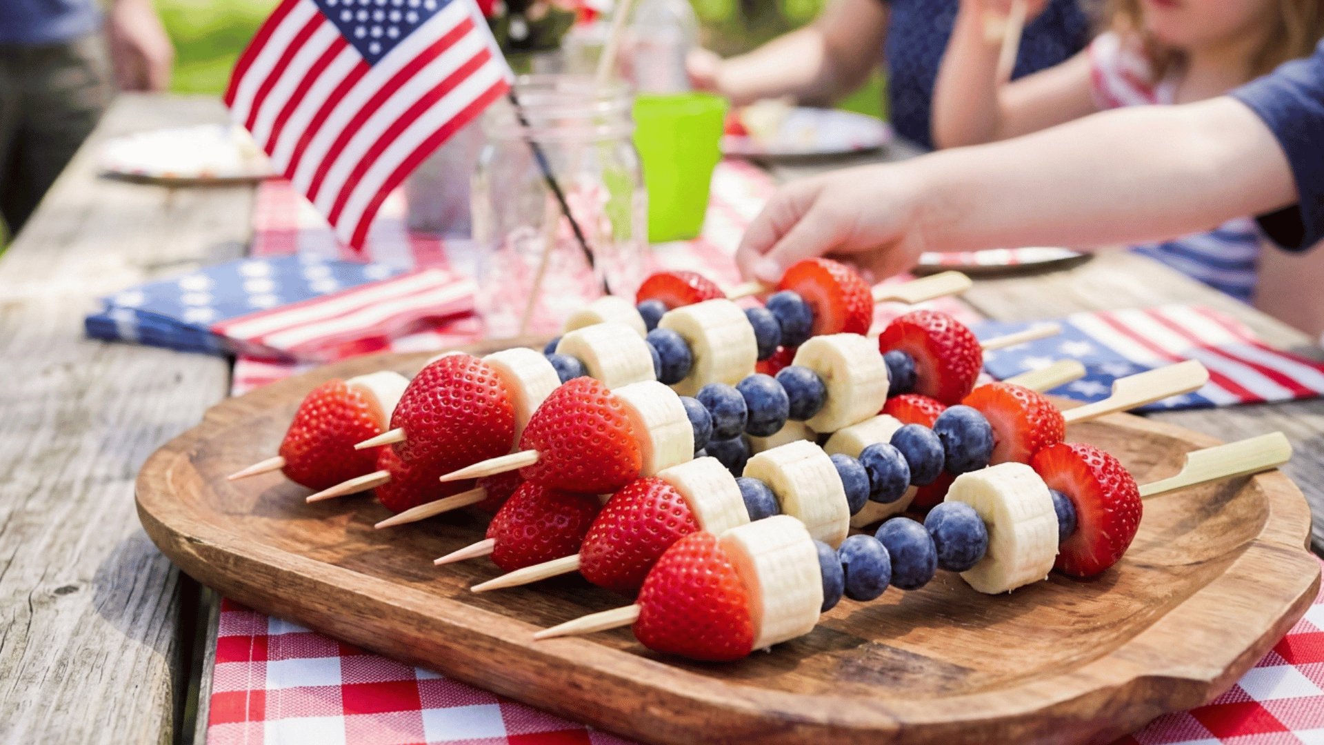 red, white, and blue fruit skewers with strawberries, bananas, and blueberries on a wooden board.
