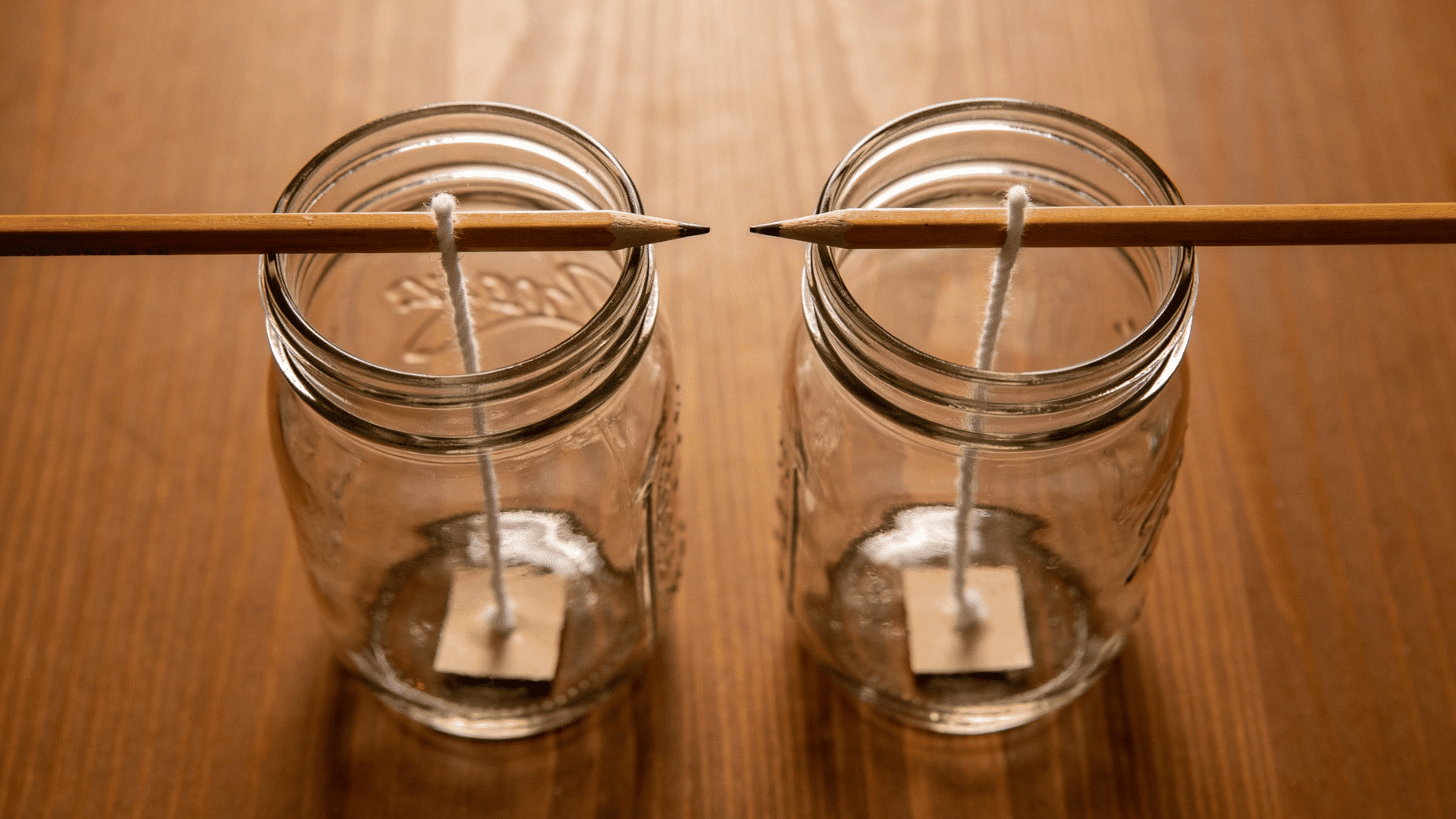 wicks centered in glass mason jars held in place by pencils laid across the top, ready for wax pouring.