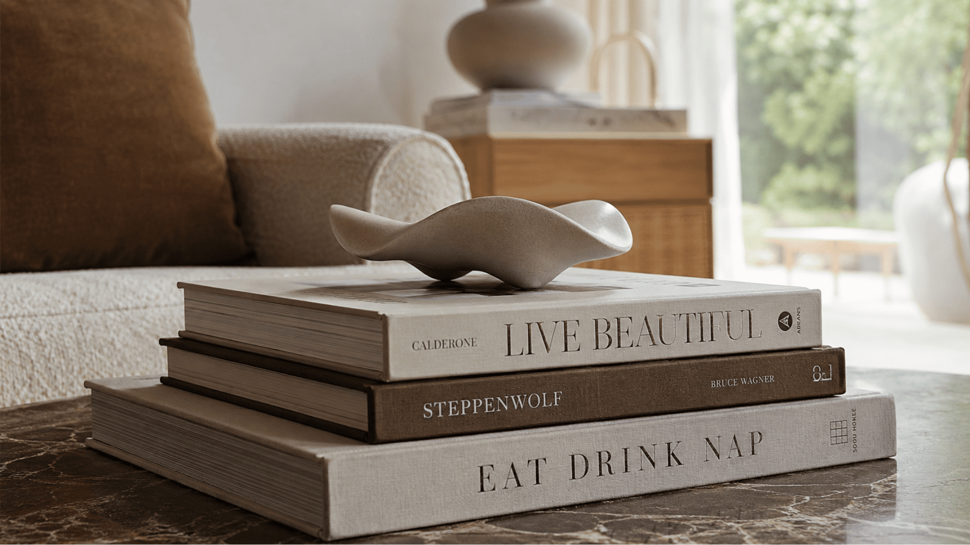 Decorative books stacked on a coffee table with a sculptural object, beside a sofa in a bright, modern living room.