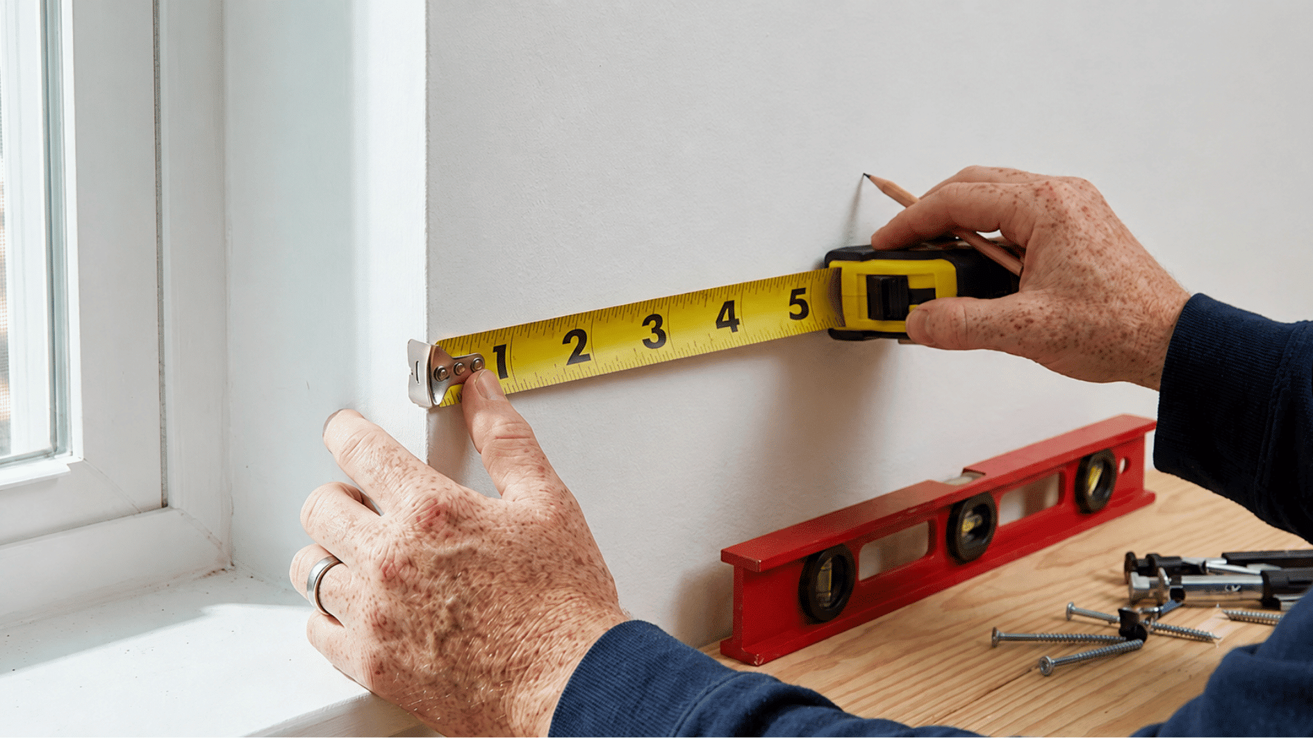 Hands measuring wall space with a tape measure and pencil to plan floating shelf placement.