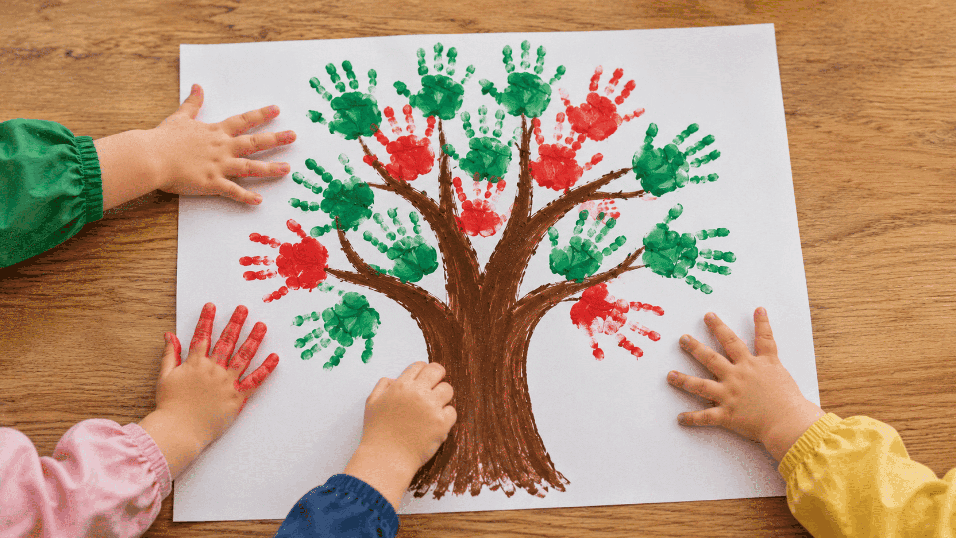 image of a handprinted apple craft handprinted apple tree visible three toddler hands painting on a white paper on a wooden table