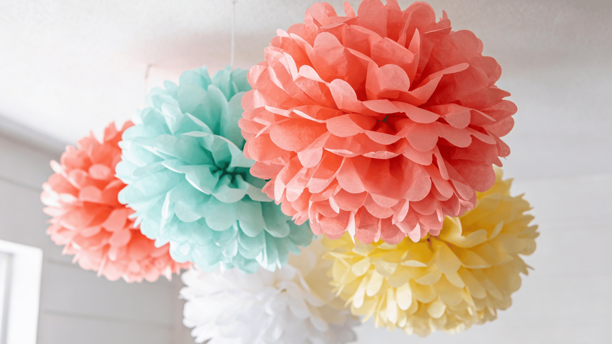 large fluffy tissue paper pom-poms in coral mint yellow and white hanging from a ceiling as birthday decor