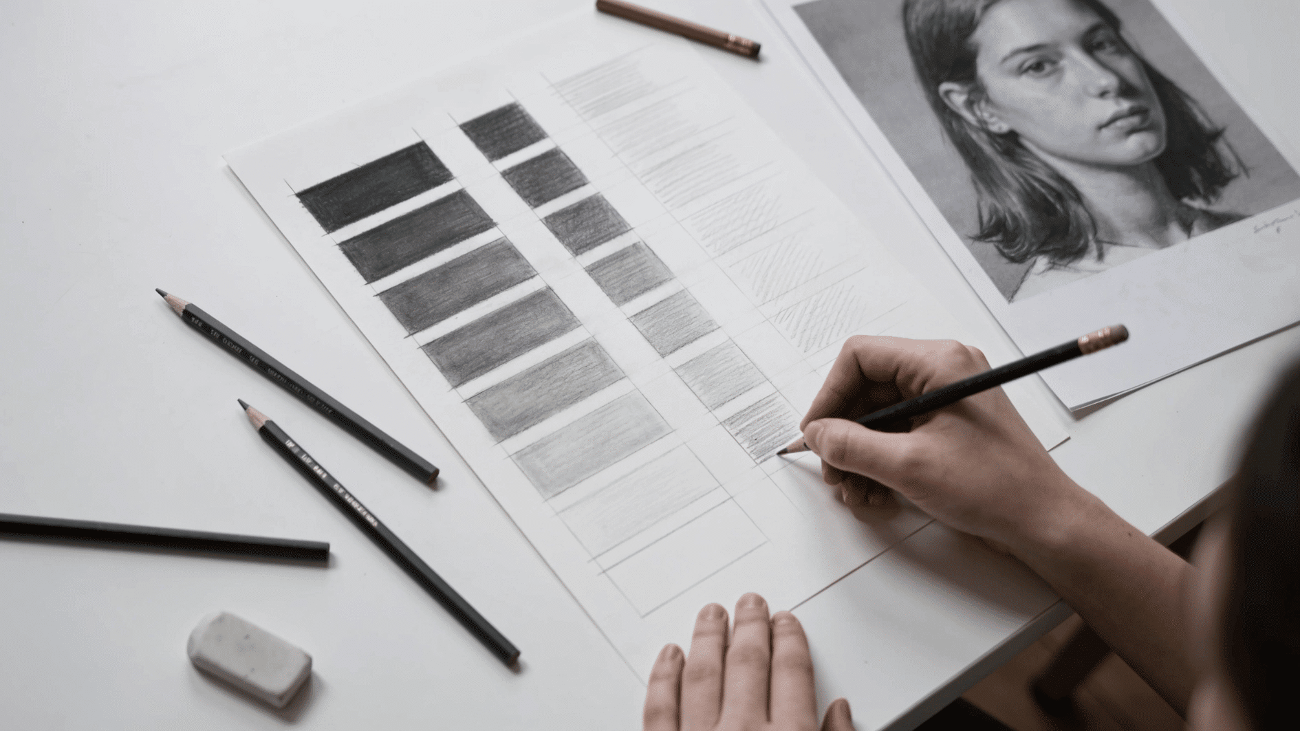 _overhead shot of a student's desk — a grayscale value scale exercise sheet mid-completion, a reference photograph nearby, pencils and an eraser scattered naturally around the page.