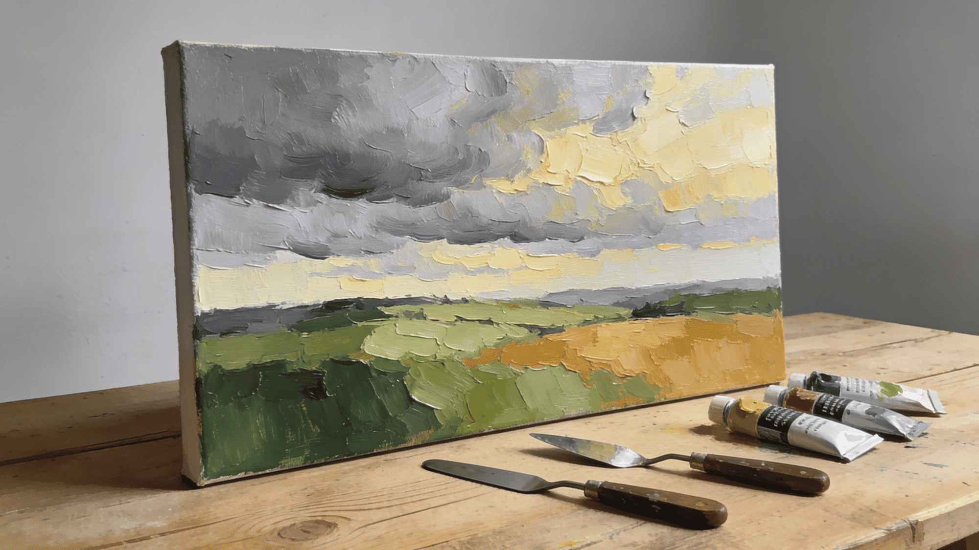 palette knife landscape textured painting on canvas with impasto sky in grey yellow and green fields on wooden worktable