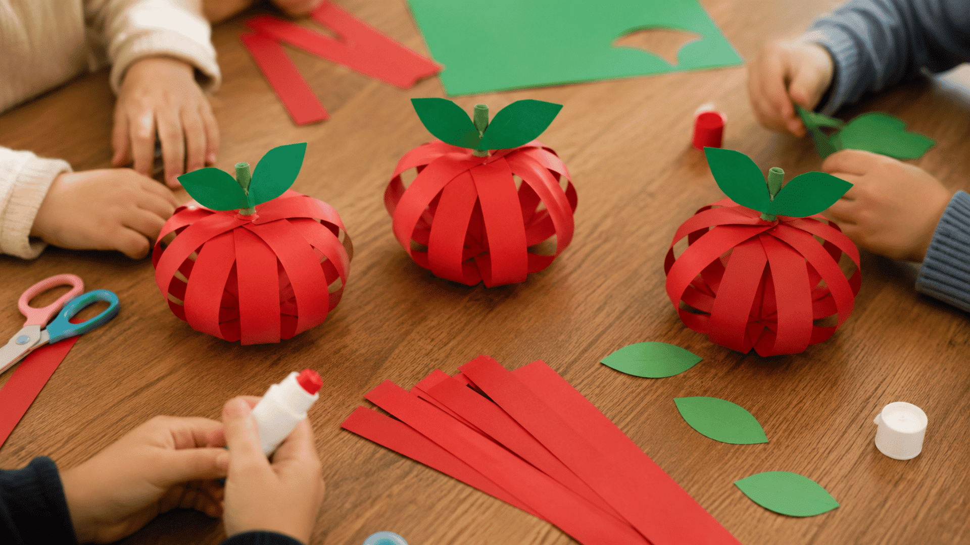 paper strip apple craft three different paper strip apples on a wooden table multiple toddlers hands visible working with craft materials on table in indoor setting