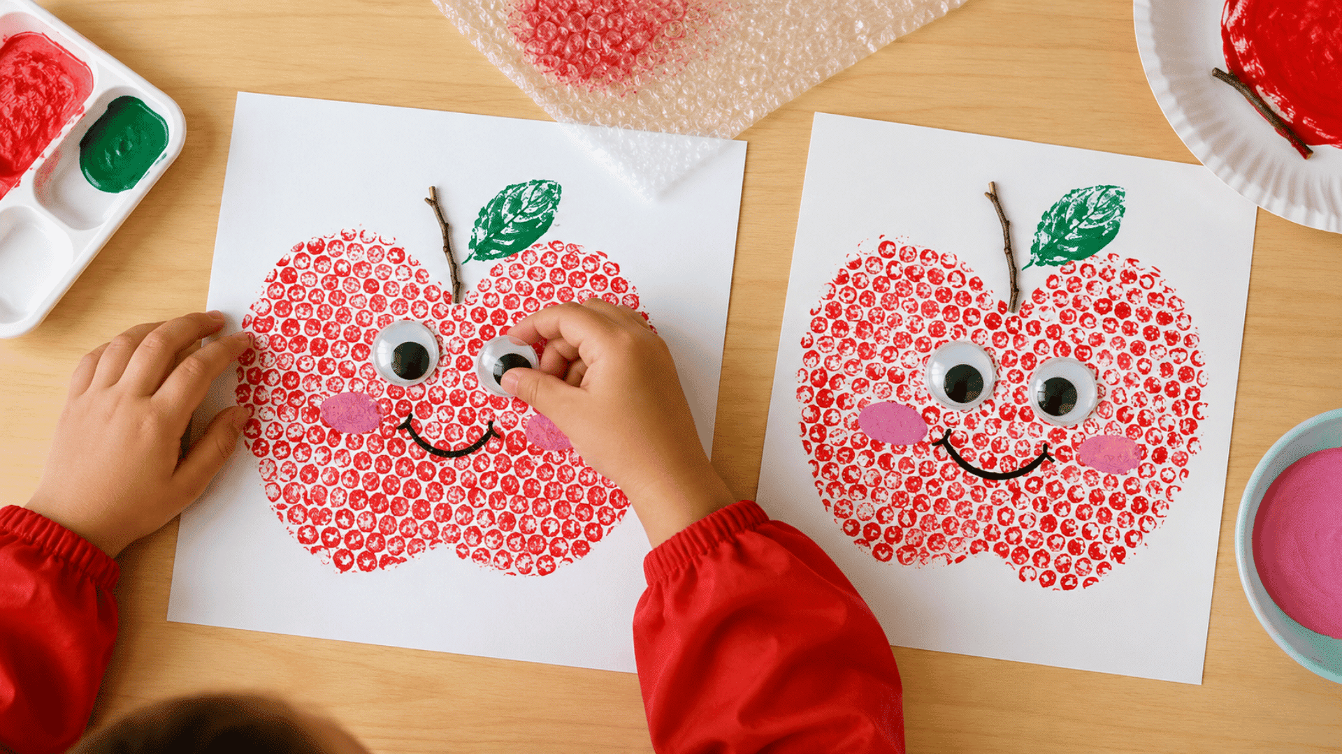 photo of a preschool craft table with bubble wrap apple print activity, large apple outlines on white paper, bubble wrap wrapped around small cardboard blocks, red washable paint on paper plates