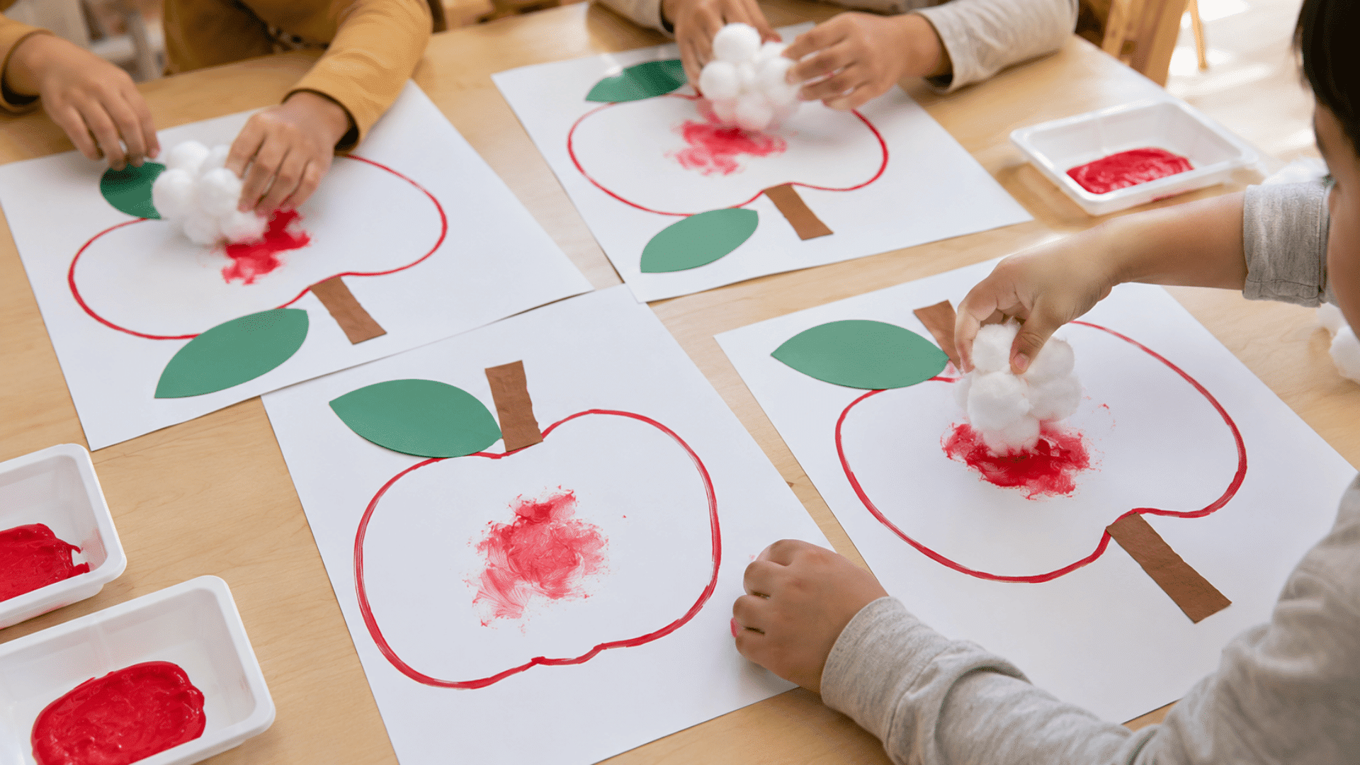 photo of a preschool craft table with cotton ball apple painting, apple outlines on white paper, red washable paint, cotton balls clipped in clothespins, green paper leaves