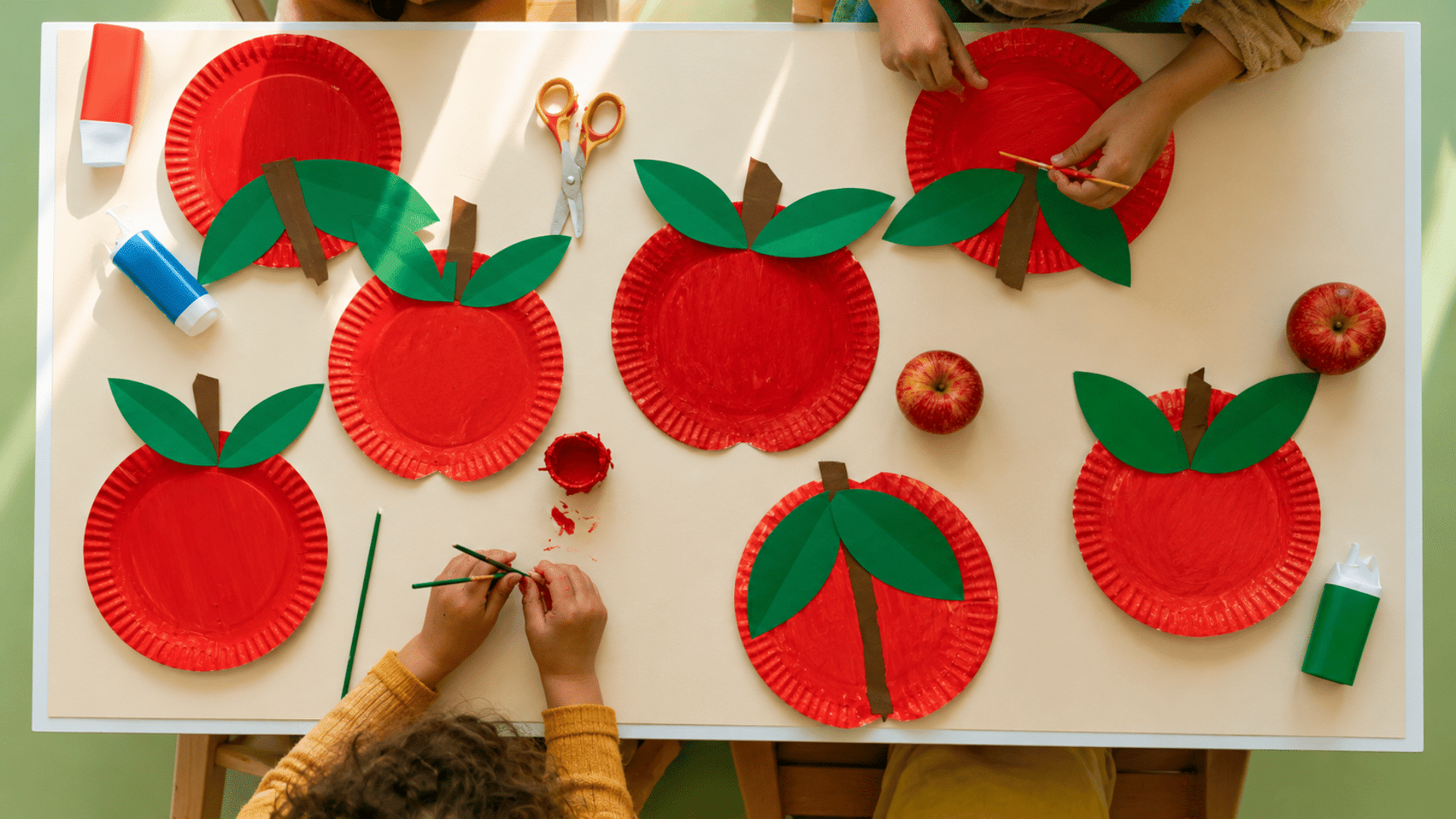photo of a preschool craft table with paper plate apple crafts, red painted paper plates, green paper leaves, brown paper stems, washable paint, glue sticks, child-safe scissors, small hands worki