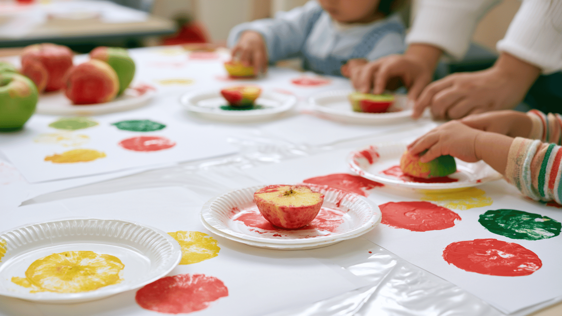 photo of a preschool craft table with real apple stamping activity, apple halves dipped in red, green, and yellow washable paint, printed apple marks on white paper