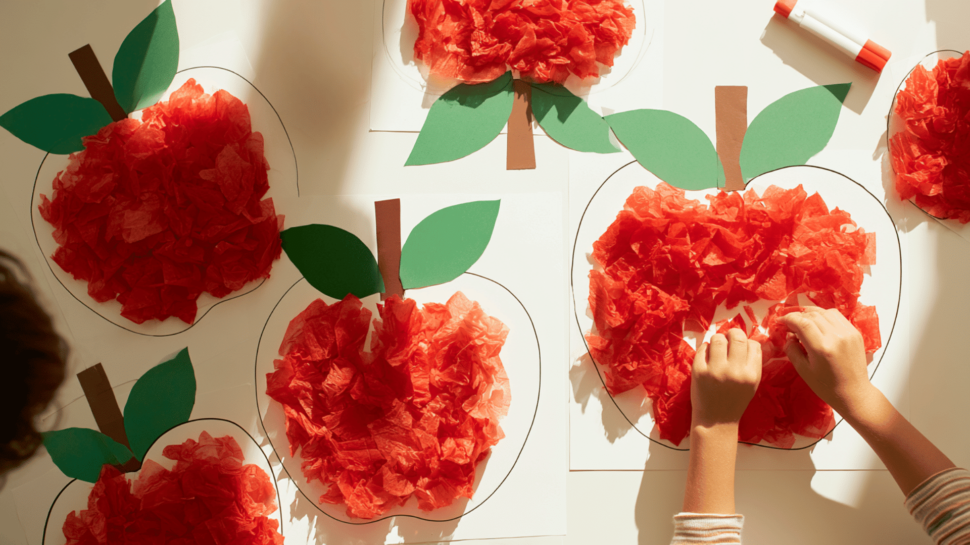 photo of a preschool craft table with tissue paper apple crafts, large apple outlines filled with crumpled red tissue paper, green paper leaves, brown stems, glue sticks nearby