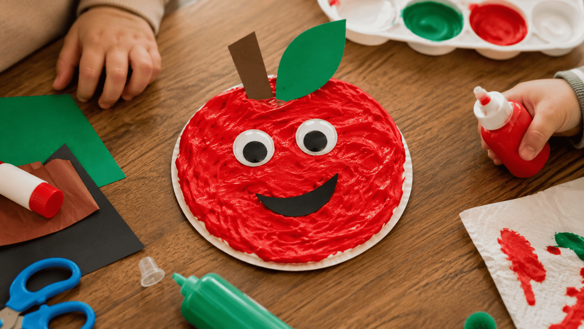 toddler apple craft idea puffy painted apple craft for display with children hands visible handling craft materials glue and paint in an indoor setting on a brown wooden table