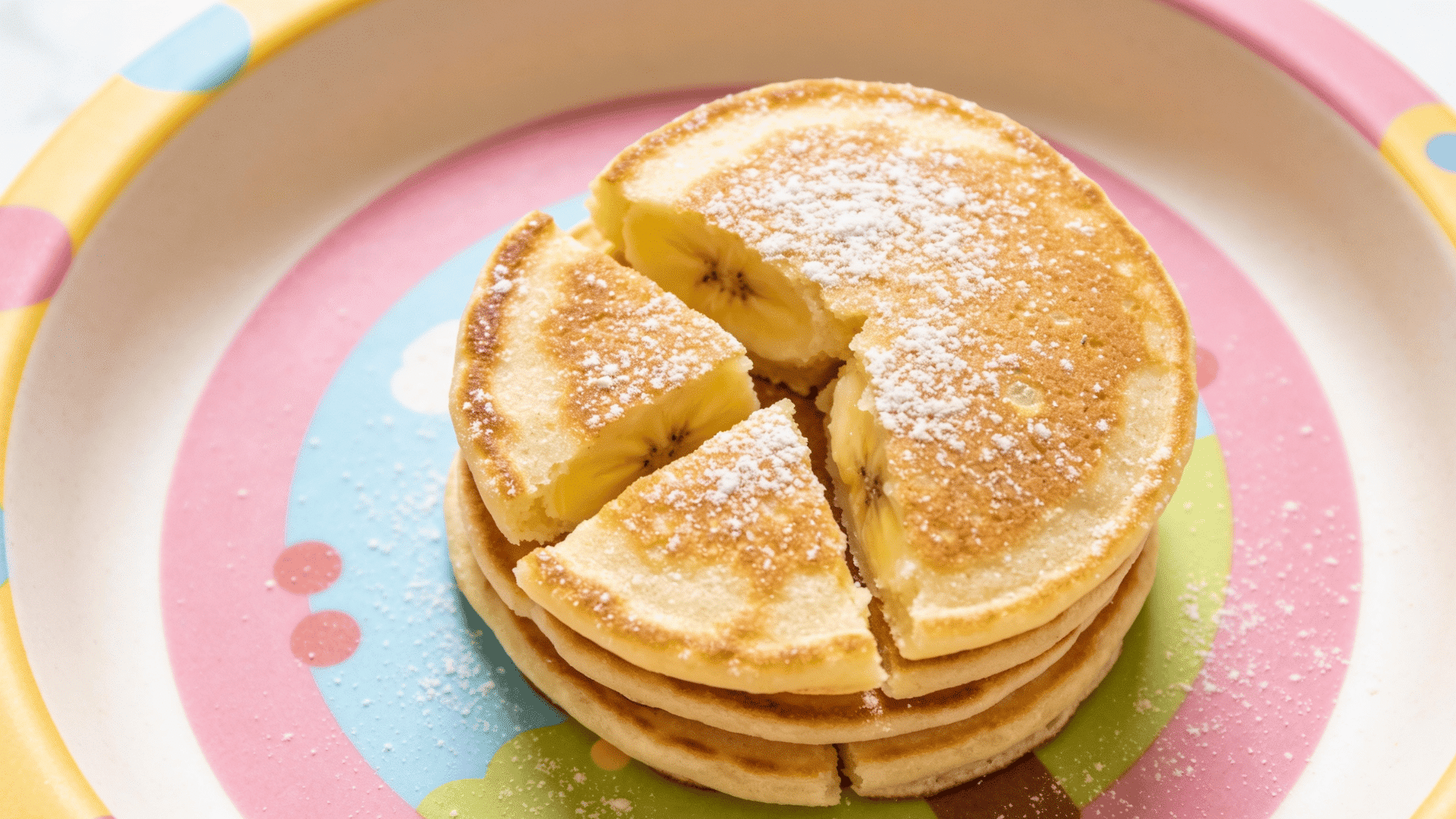 A short stack of small soft banana pancakes cut into toddler-friendly pieces on a pastel plate, dusting of nothing added, cheerful bright kitchen, overhead view.