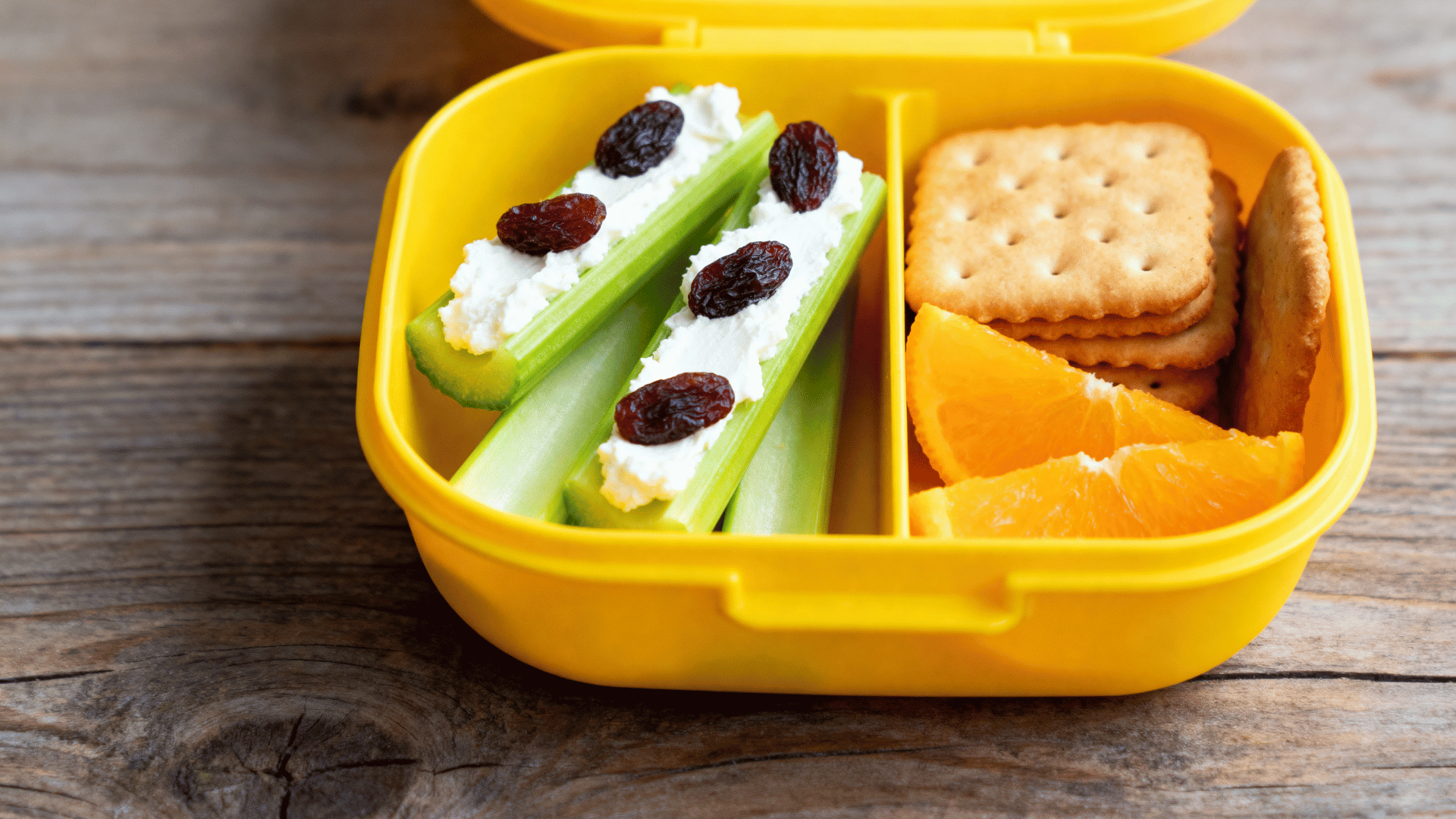 celery sticks with cream cheese and raisins, crackers, and orange wedges in a buttercup-yellow lunchbox