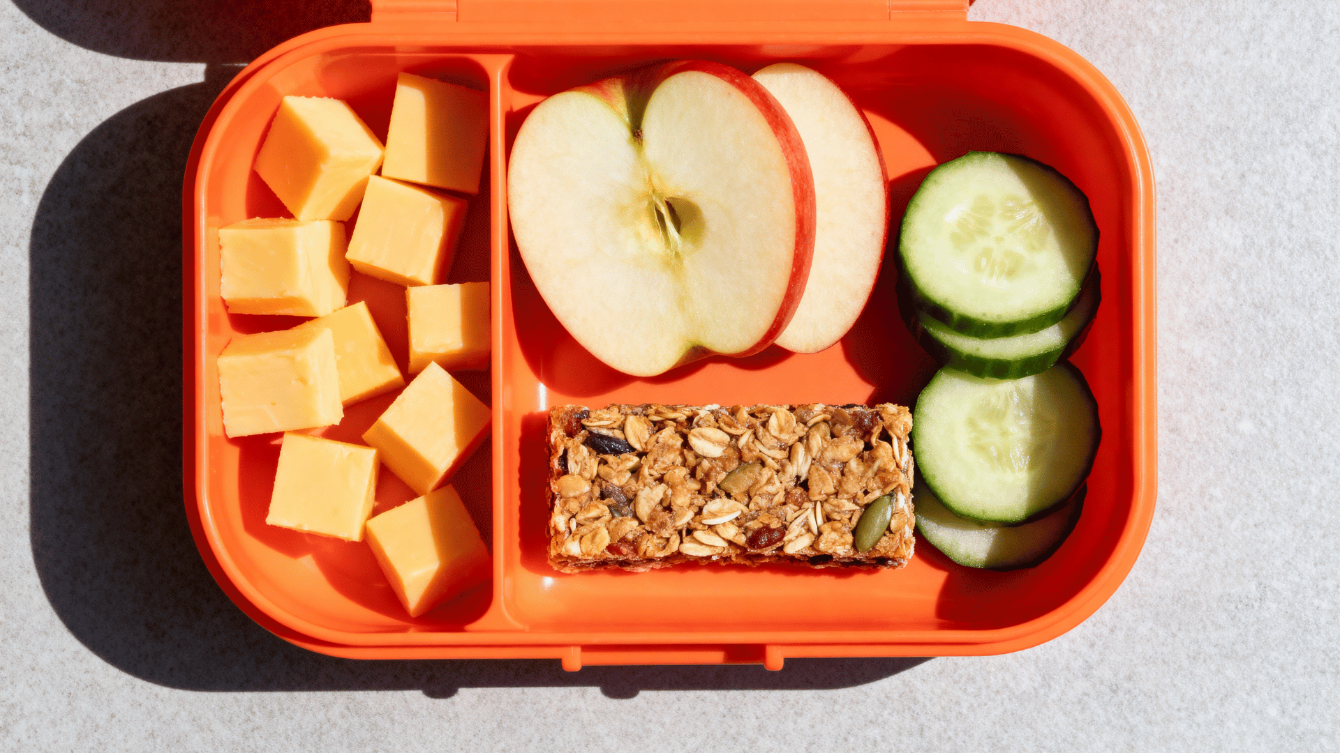 cheese cubes, apple slices, granola bar, and cucumber in a coral-orange silicone lunchbox
