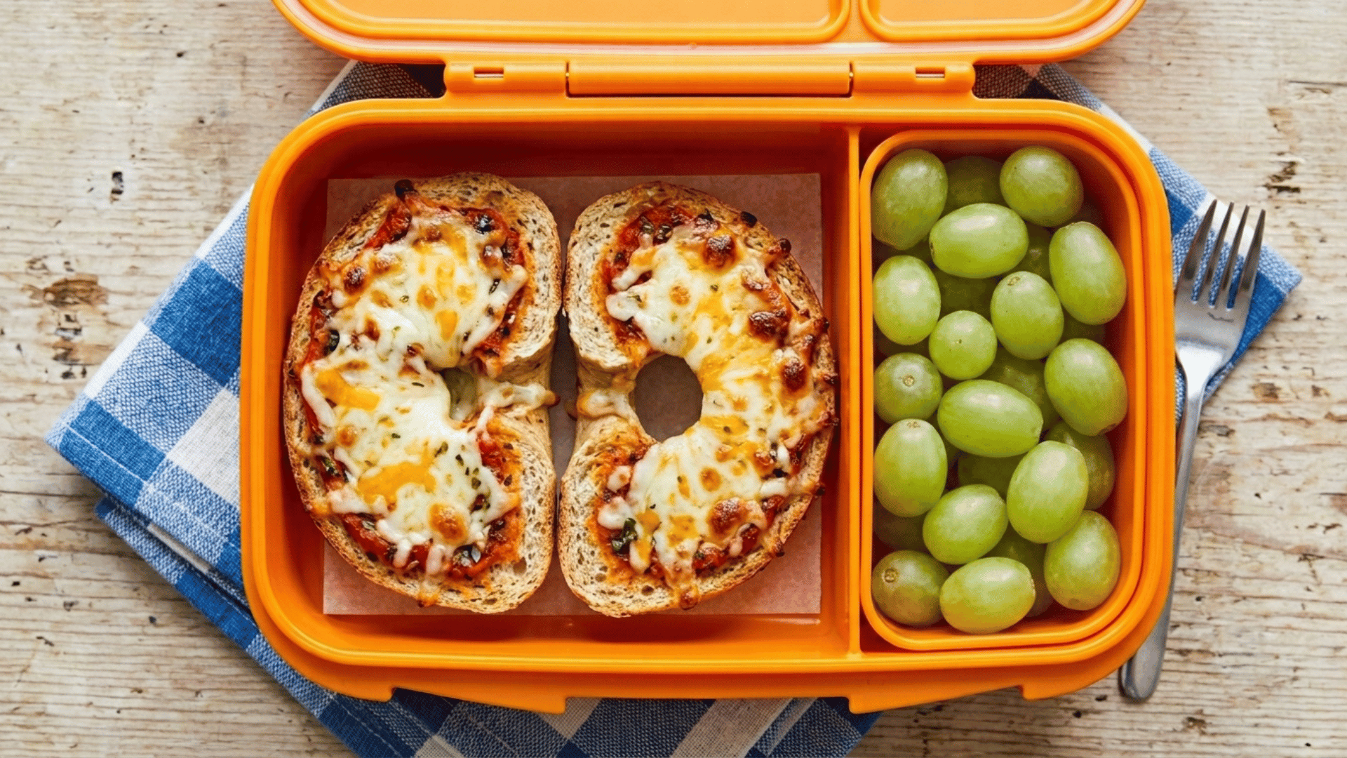 mini cheese pizza bagels packed in an orange plastic lunchbox with green grapes
