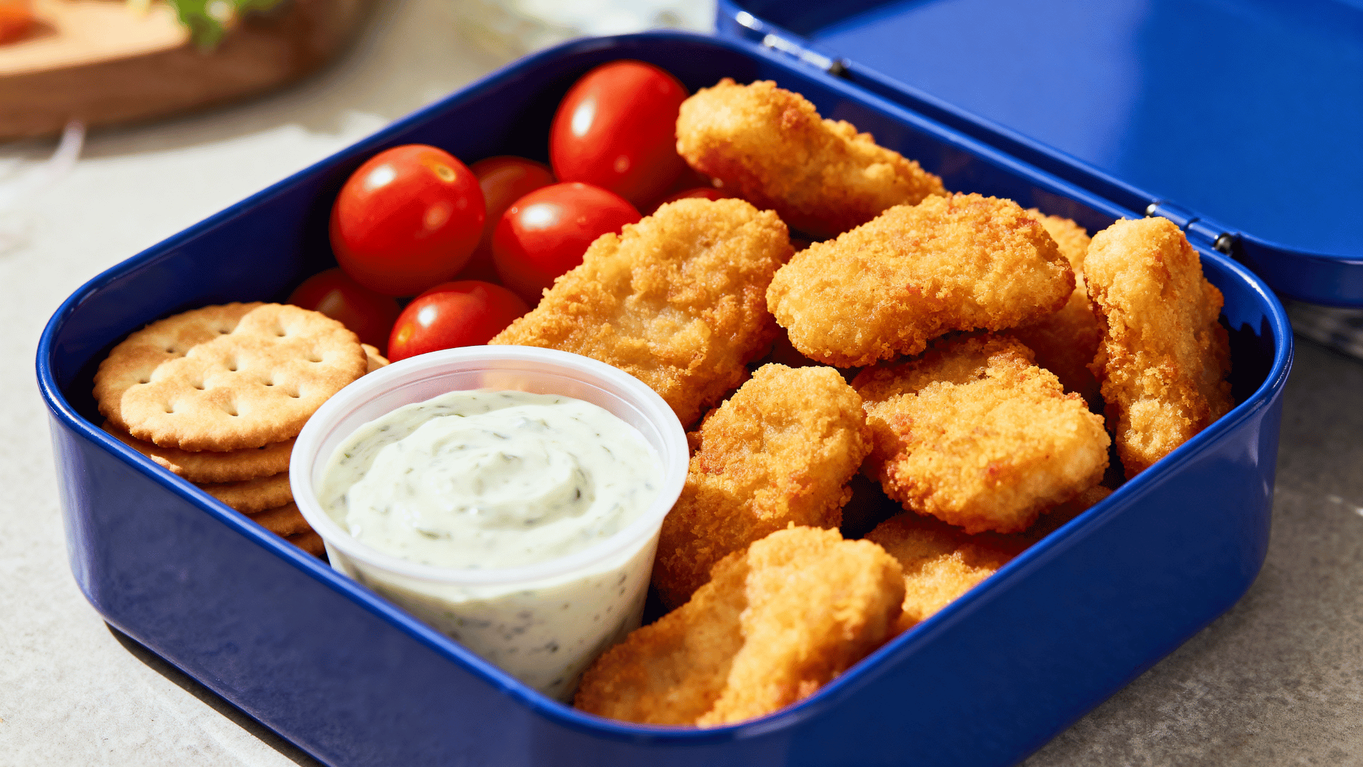chicken nuggets, ranch dip, cherry tomatoes, and crackers in a cobalt-blue enamel lunch tin