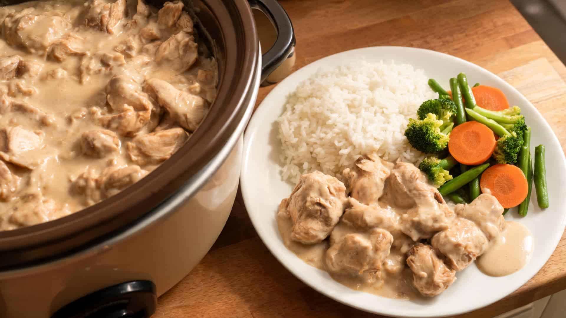 creamy chicken served with rice and steamed vegetables on a white plate beside a slow cooker on a wooden table