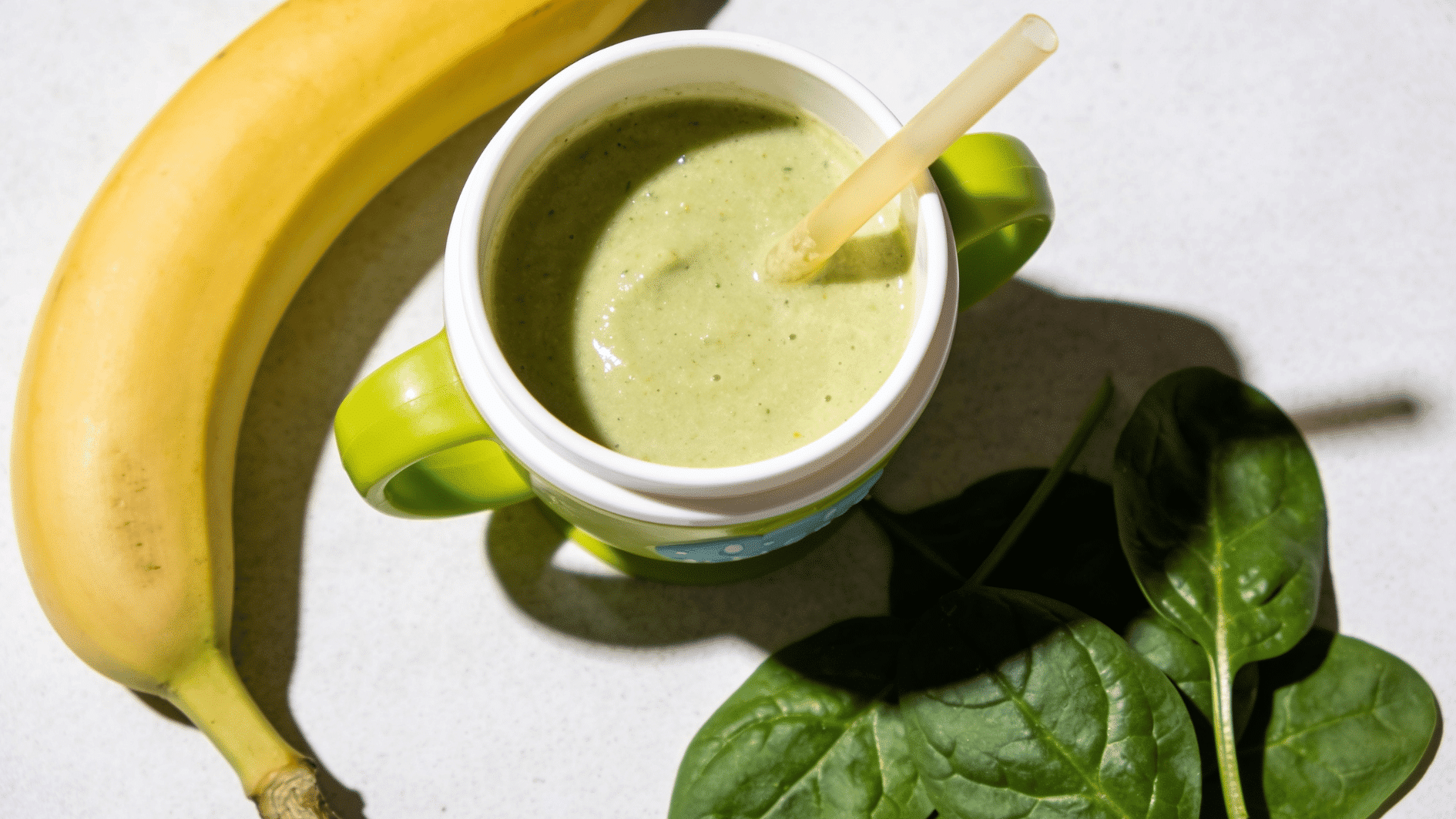 creamy green-tinged fruit smoothie in a small toddler cup with a straw, fresh banana and spinach leaves beside it,