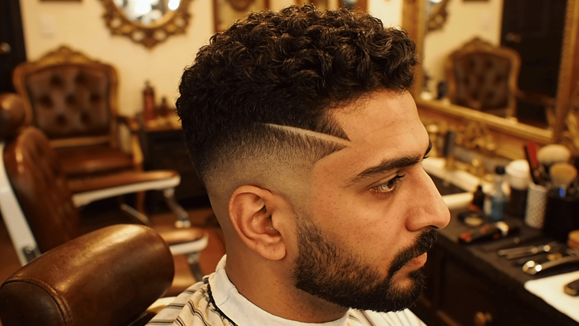curly textured fade haircut with shaved line design and beard styling in a warm toned barbershop setting