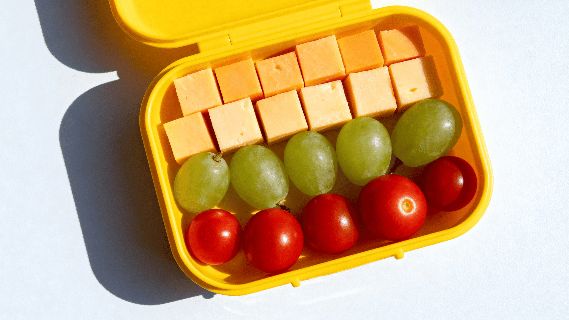 cheese, grapes, and tomatoes arranged in rows in a yellow kids lunchbox