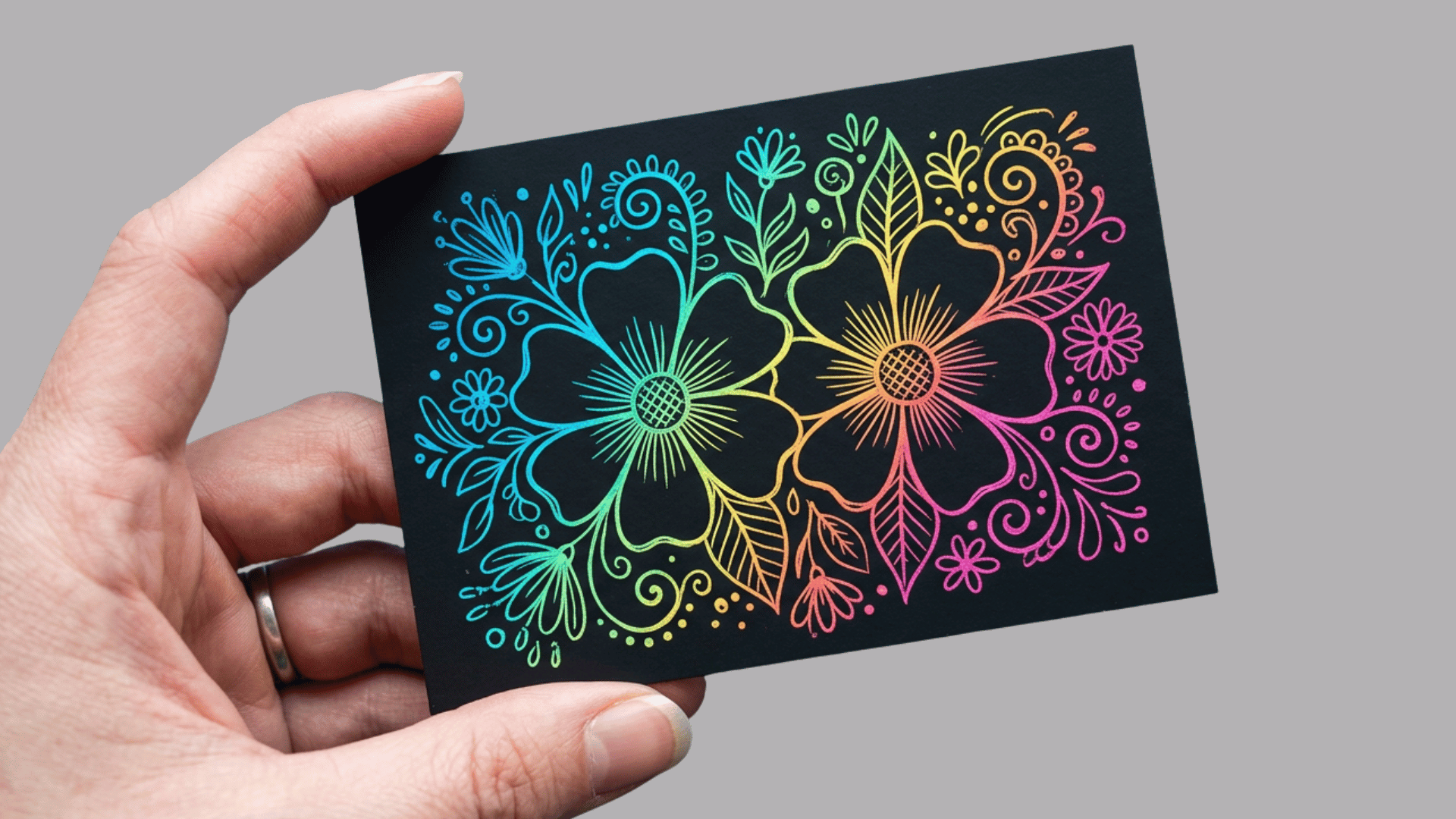 hand holding a black greeting card with vibrant rainbow neon floral illustration featuring two large flowers and intricate leaves in cyan, green, yellow and pink