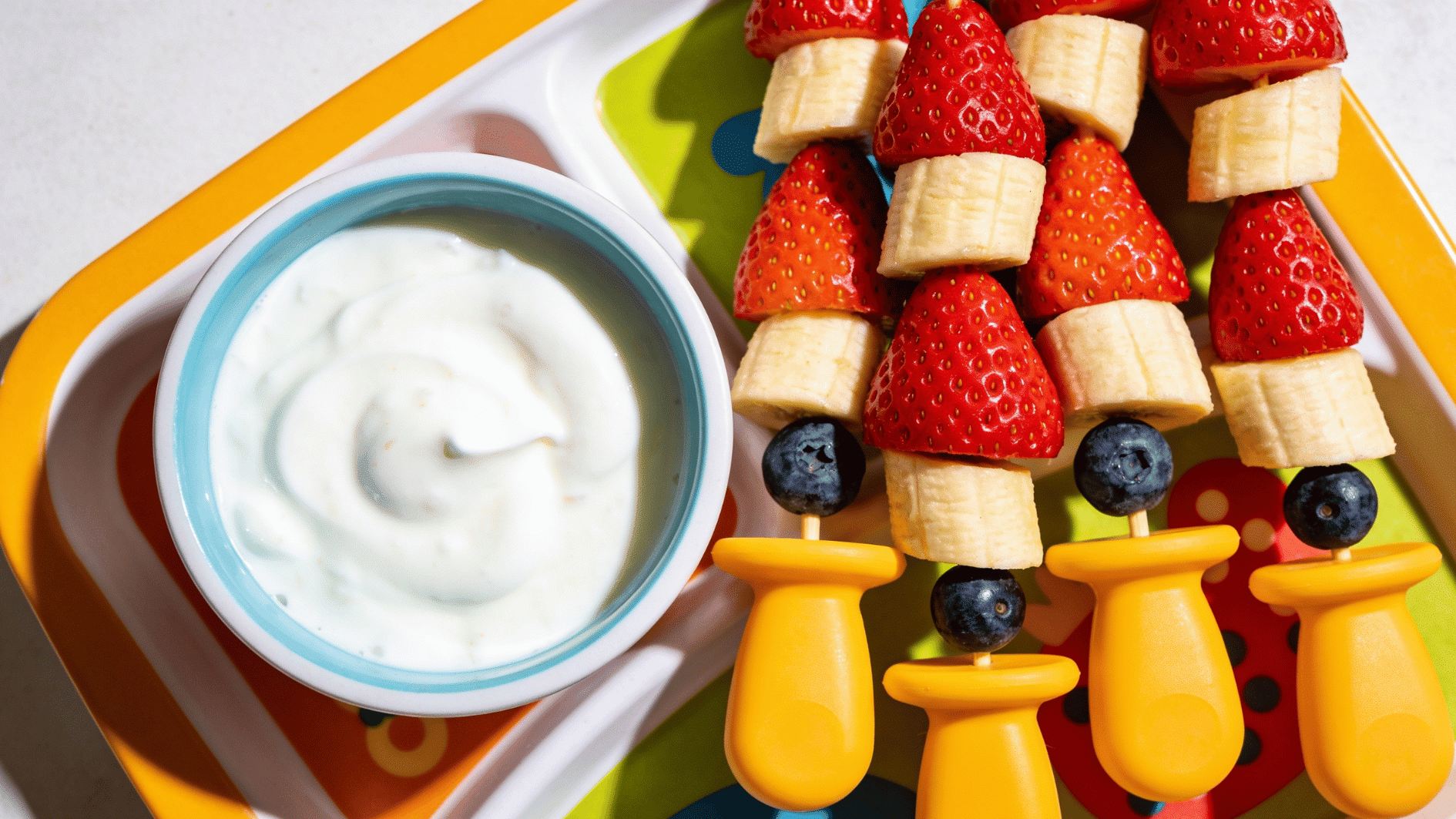fruit pieces on short blunt toddler-safe skewers beside a small bowl of yogurt for dipping