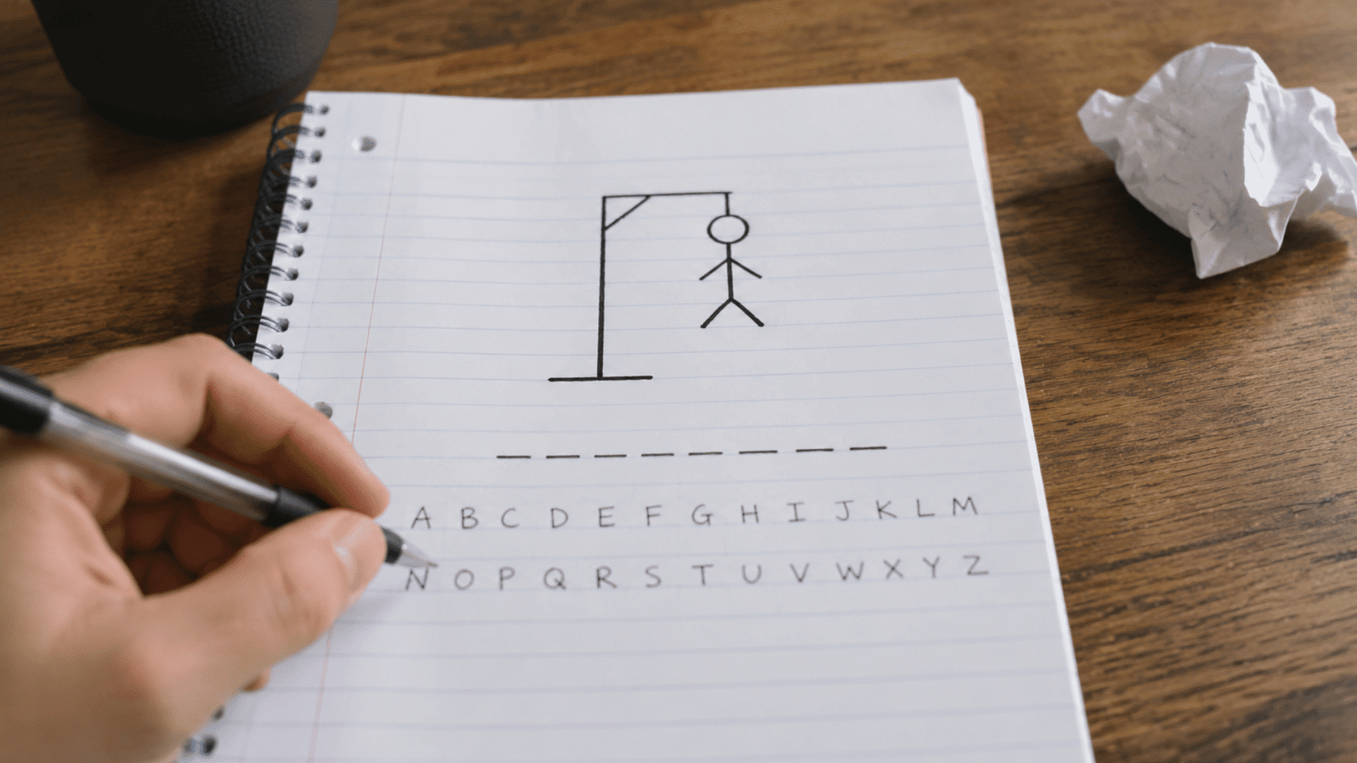 hand holding a pen playing hangman on a spiral notebook with a drawing of a stick figure and letters of the alphabet