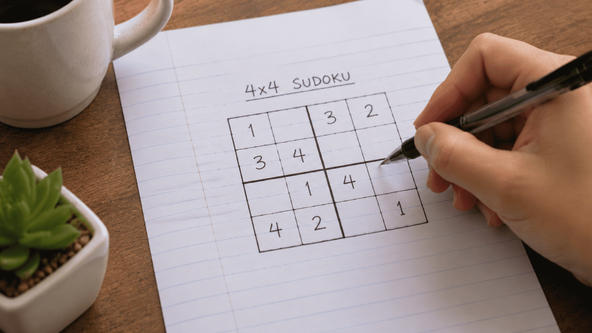 hand holding a pen solving a 4x4 sudoku puzzle drawn on a sheet of lined paper
