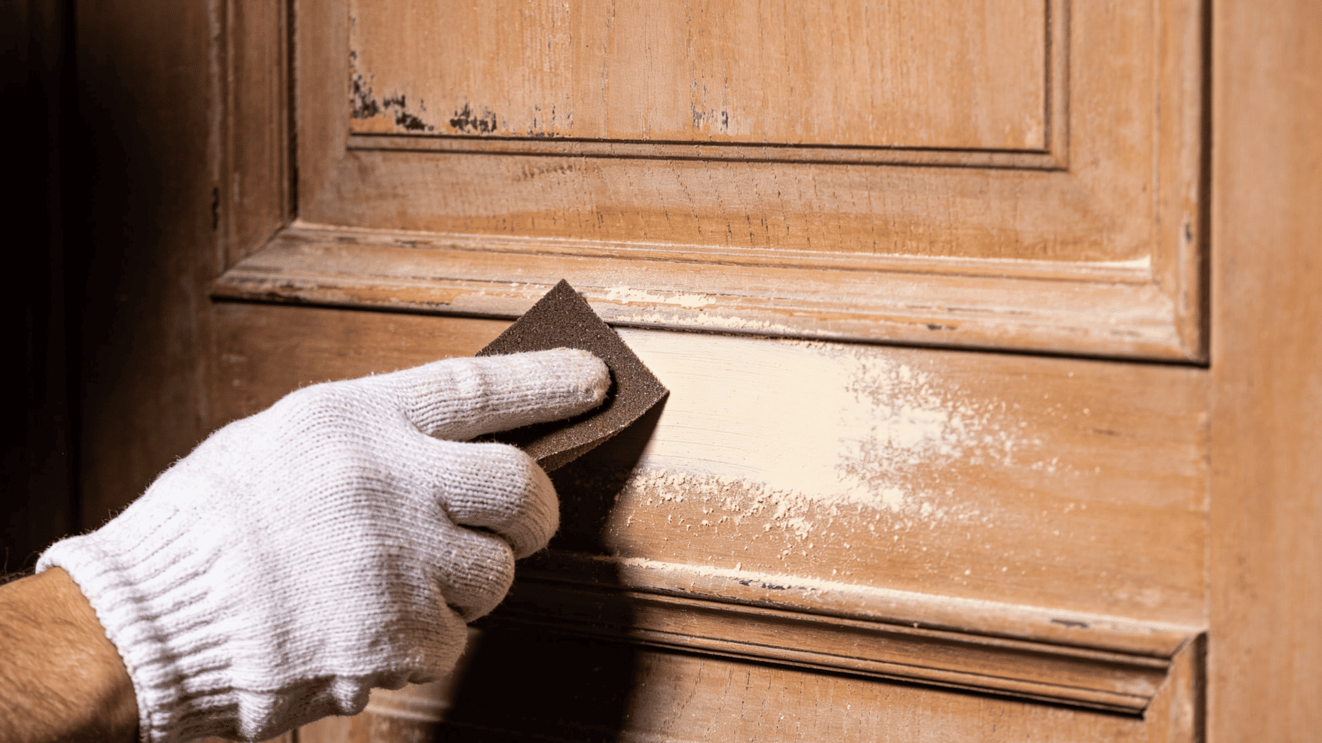 hand wearing glove sanding a wooden door surface to remove old paint and prepare the area for refinishing work