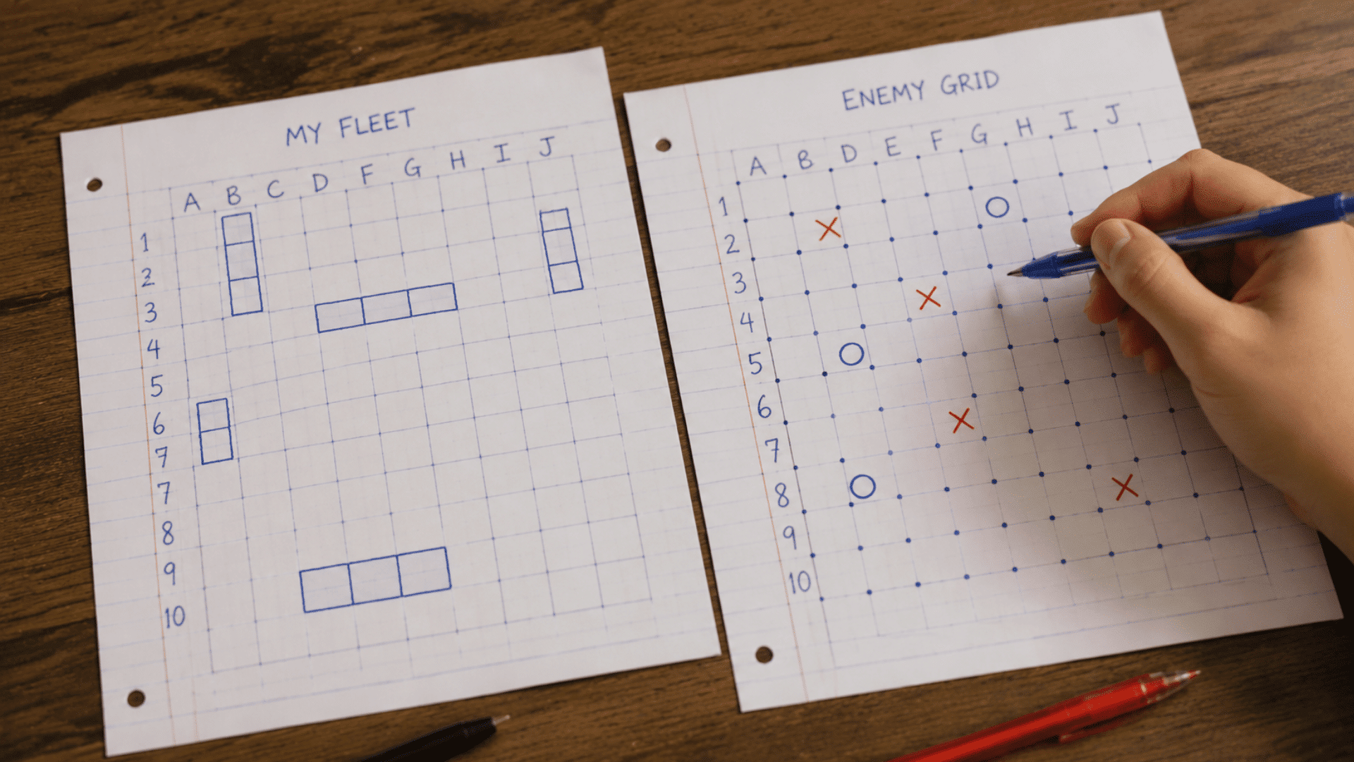 hand with a blue pen playing battleship on paper sheets labeled my fleet and enemy grid