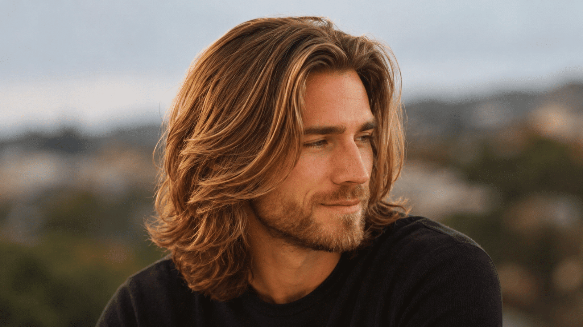 man with long blond layered hair and beard wearing a dark shirt in soft golden outdoor light