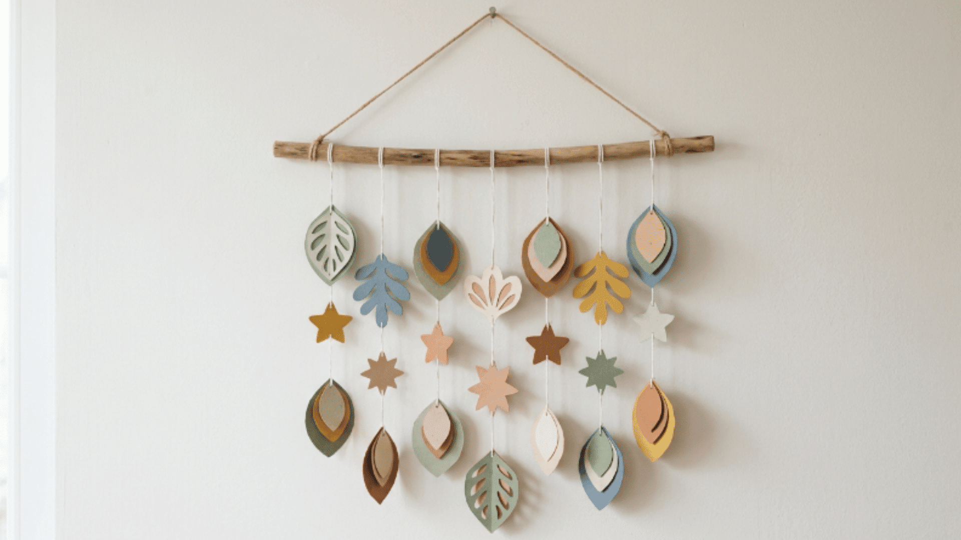 paper wall hanging with cut shapes attached to strings hanging from a wooden stick against a plain wall