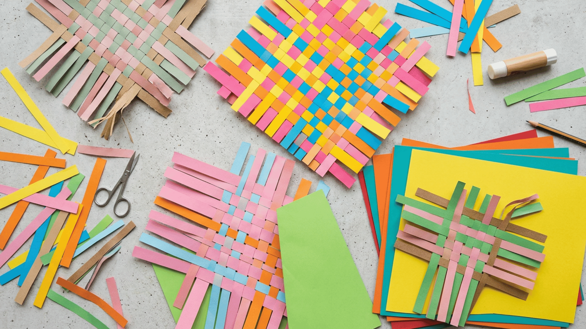 colorful paper weaving craft project on gray surface with vibrant woven mats made of pink, yellow, blue and green strips, surrounded by scissors, glue and loose paper