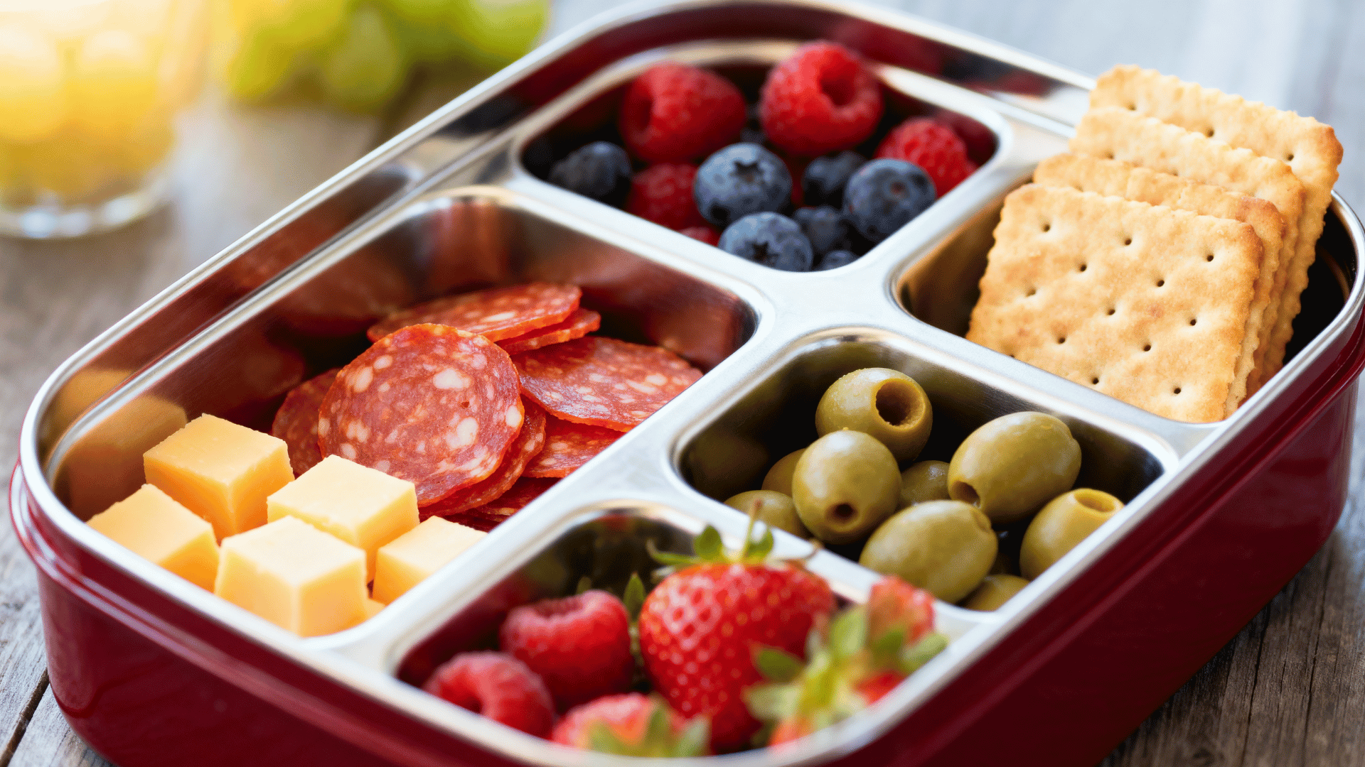 pepperoni slices, cheese cubes, olives, crackers, and berries in a maroon-red steel lunchbox
