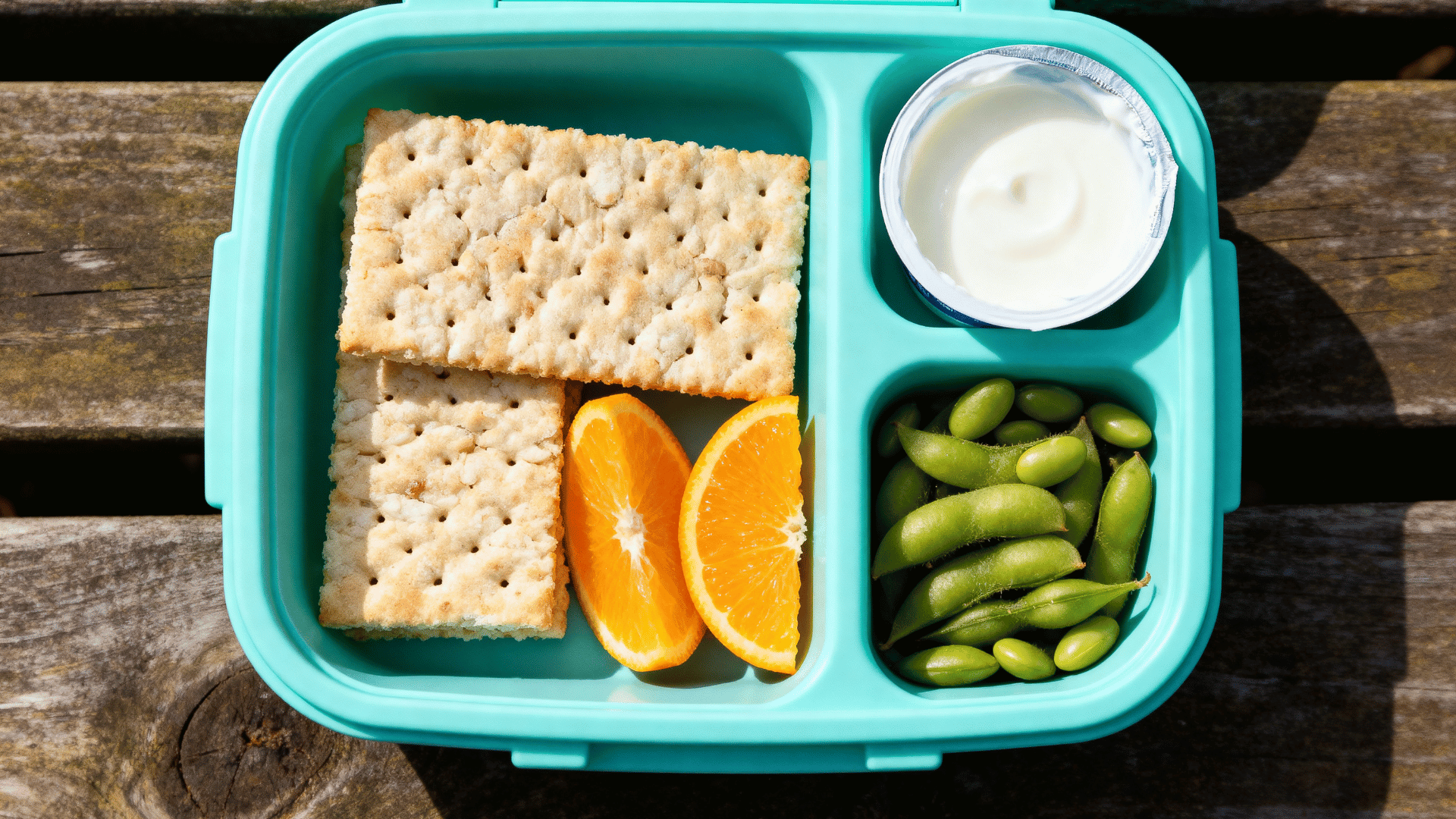 rice crackers, edamame, orange slices, and yogurt cup in an aqua-blue silicone lunchbox