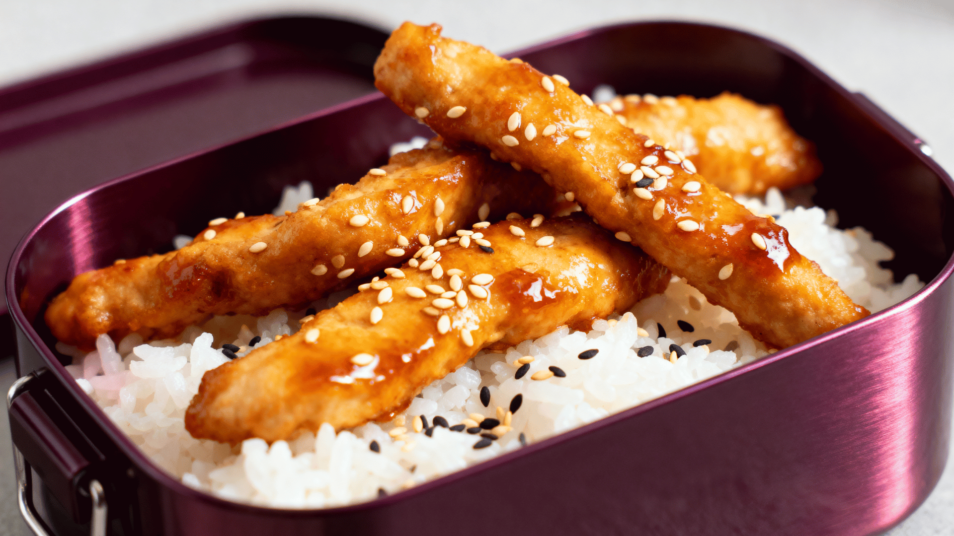 sesame chicken strips over rice in a deep plum-purple steel bento box
