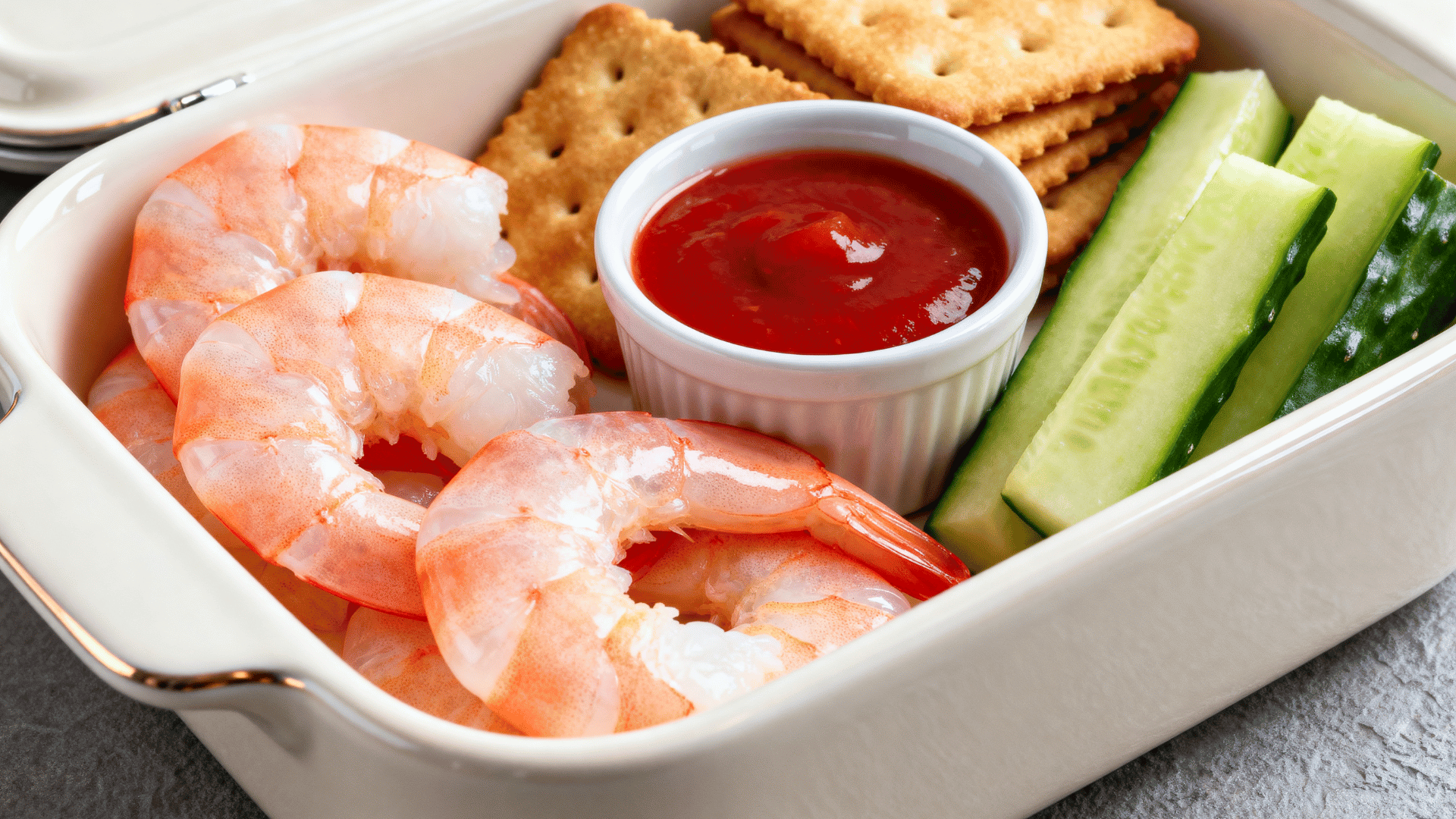 chilled shrimp, cocktail sauce, cucumber sticks, and crackers in a pearl-white ceramic lunchbox