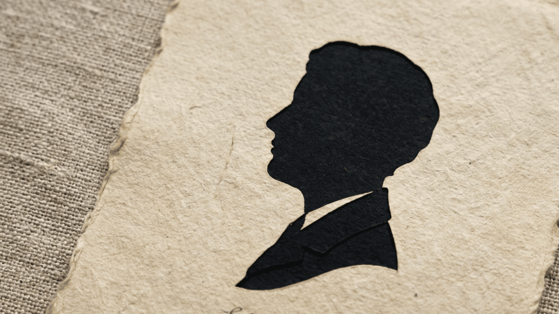 black paper silhouette portrait of a person in profile on aged handmade paper with handwritten name below, resting on textured fabric