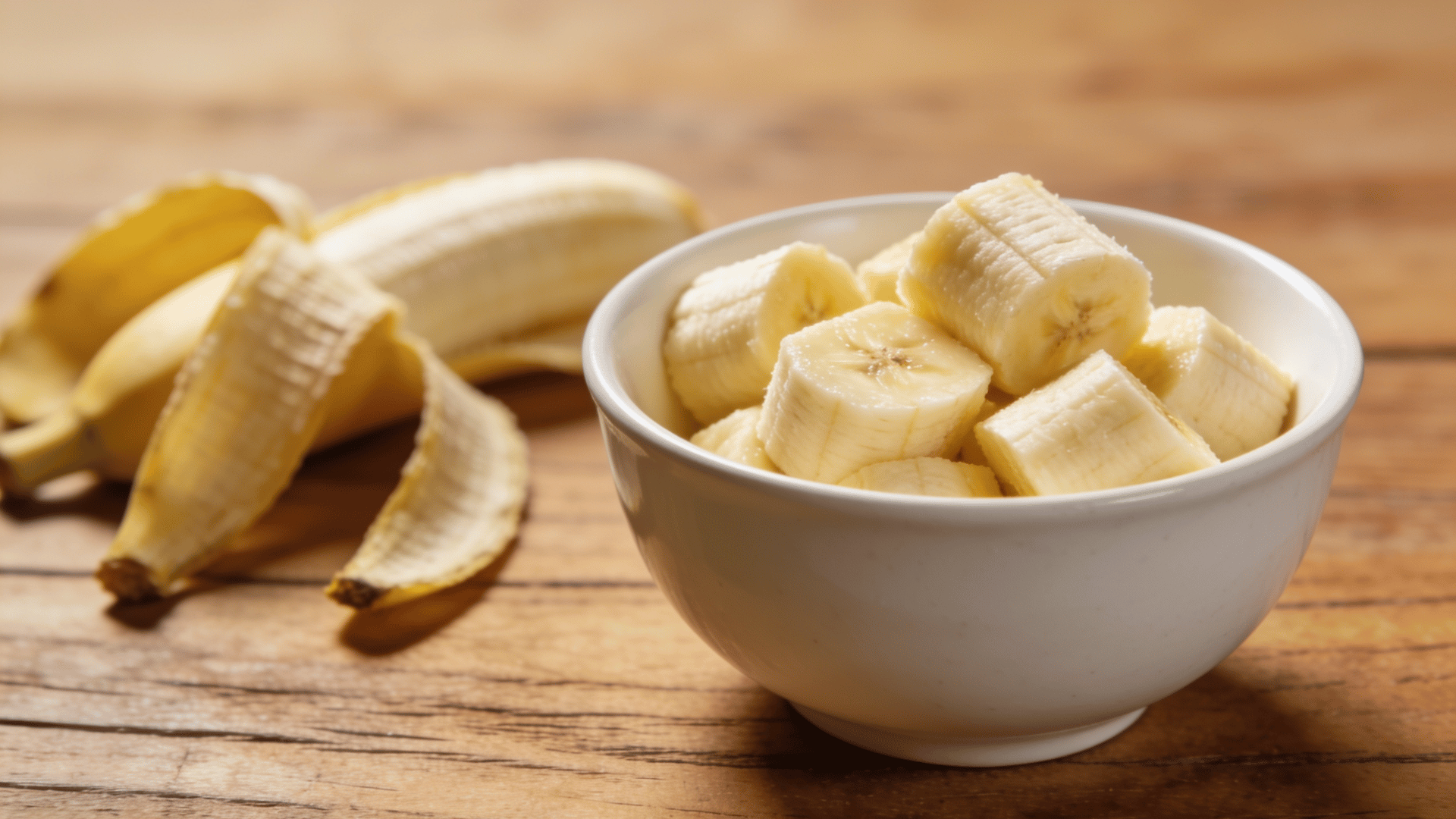 sliced banana pieces in white bowl on wooden kitchen table with banana peel in background warm natural light close views