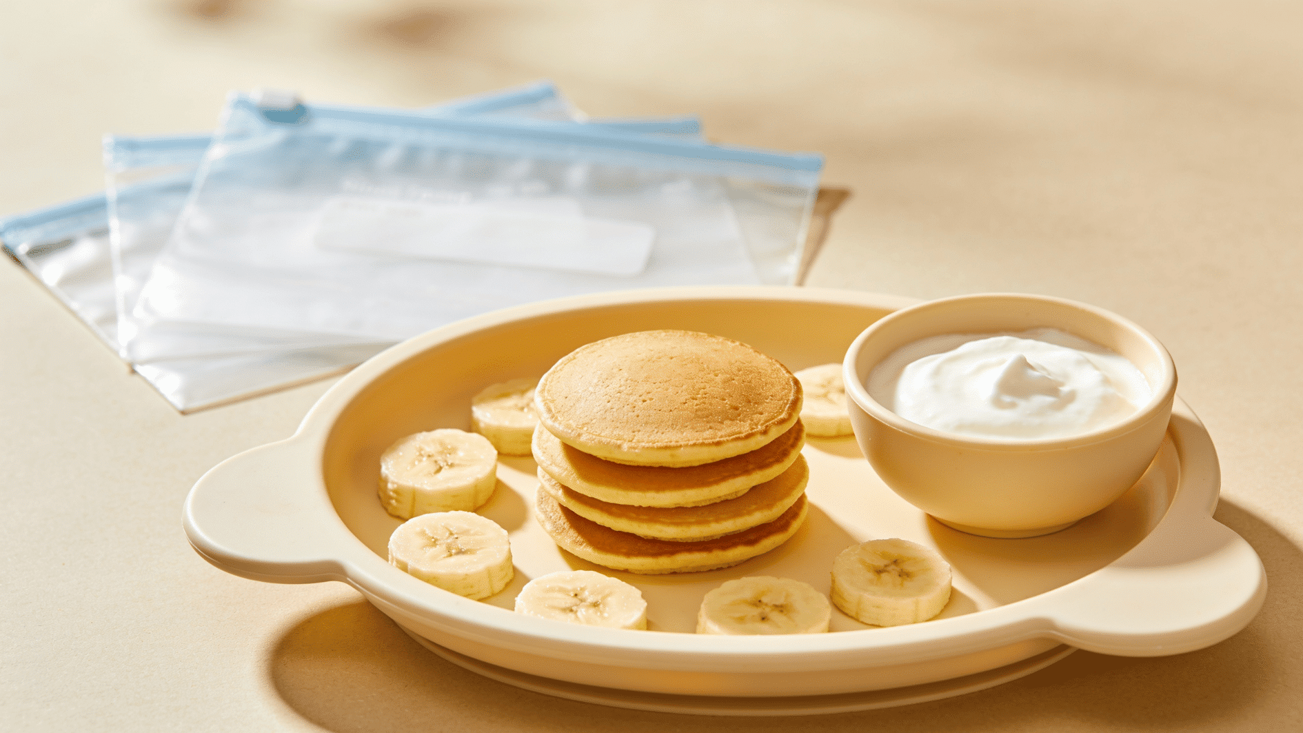 small toddler-sized pancakes stacked on a child-safe plate with banana slices, yogurt dip, and a few freezer storage bags in the background
